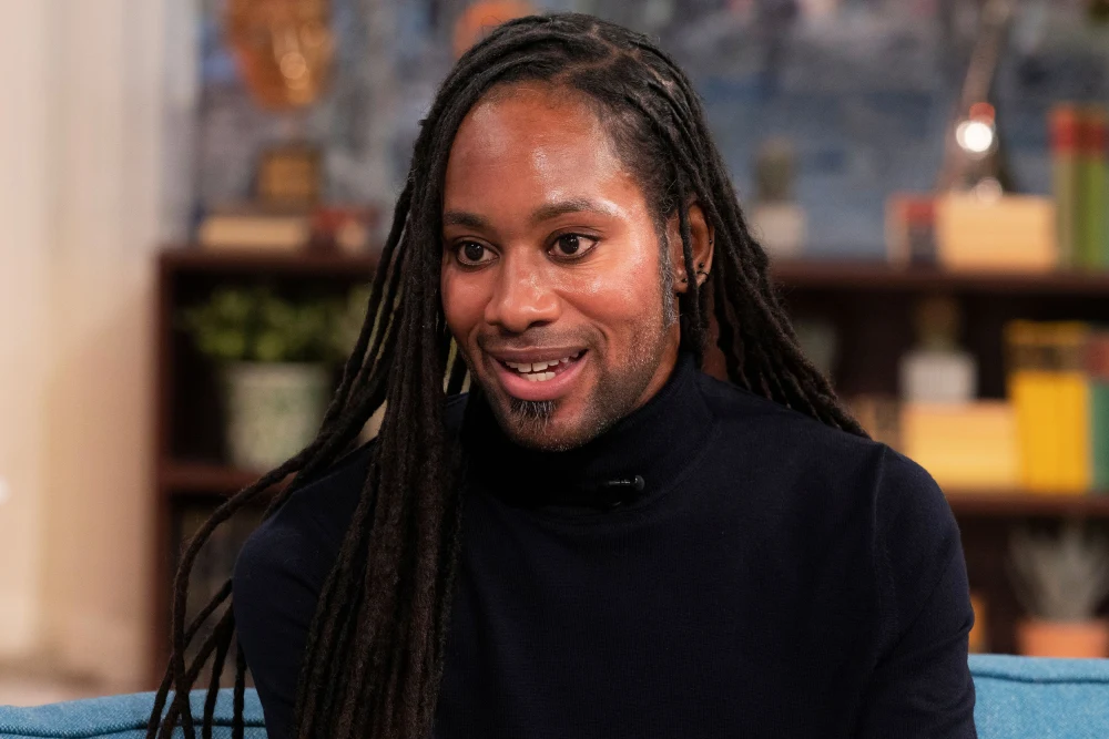 Jason Arday, wearing a black turtleneck, seated and smiling during an interview.