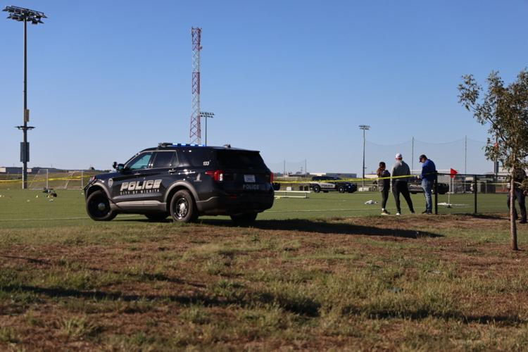 A police SUV parked on a grass soccer field with people standing in the background under a clear sky.