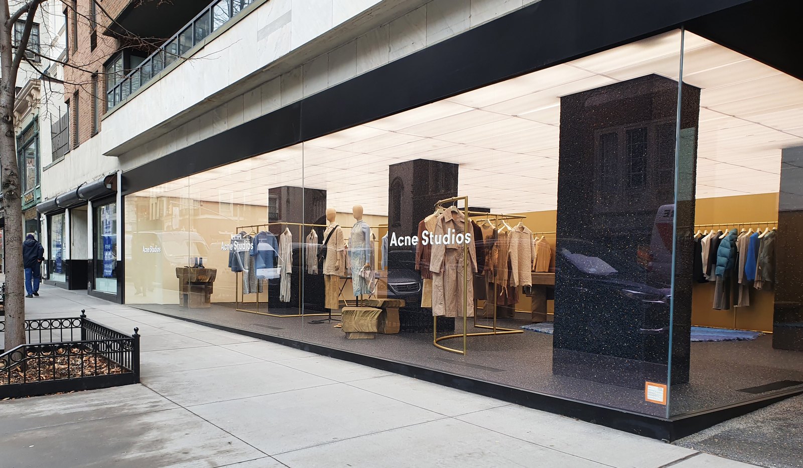 A corner Acne Studios boutique with floor-to-ceiling glass windows, showing racks of coats and knitwear and a mannequin display on a city sidewalk.