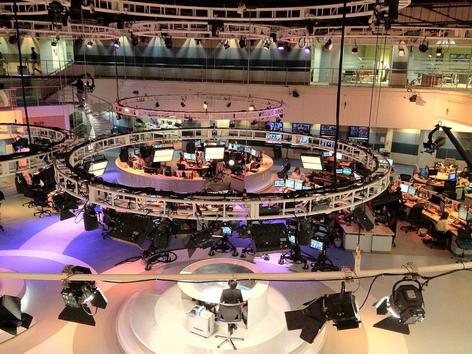Overhead view of the circular Al Jazeera English newsroom in Doha, with rows of journalists' desks and screens around a central studio.