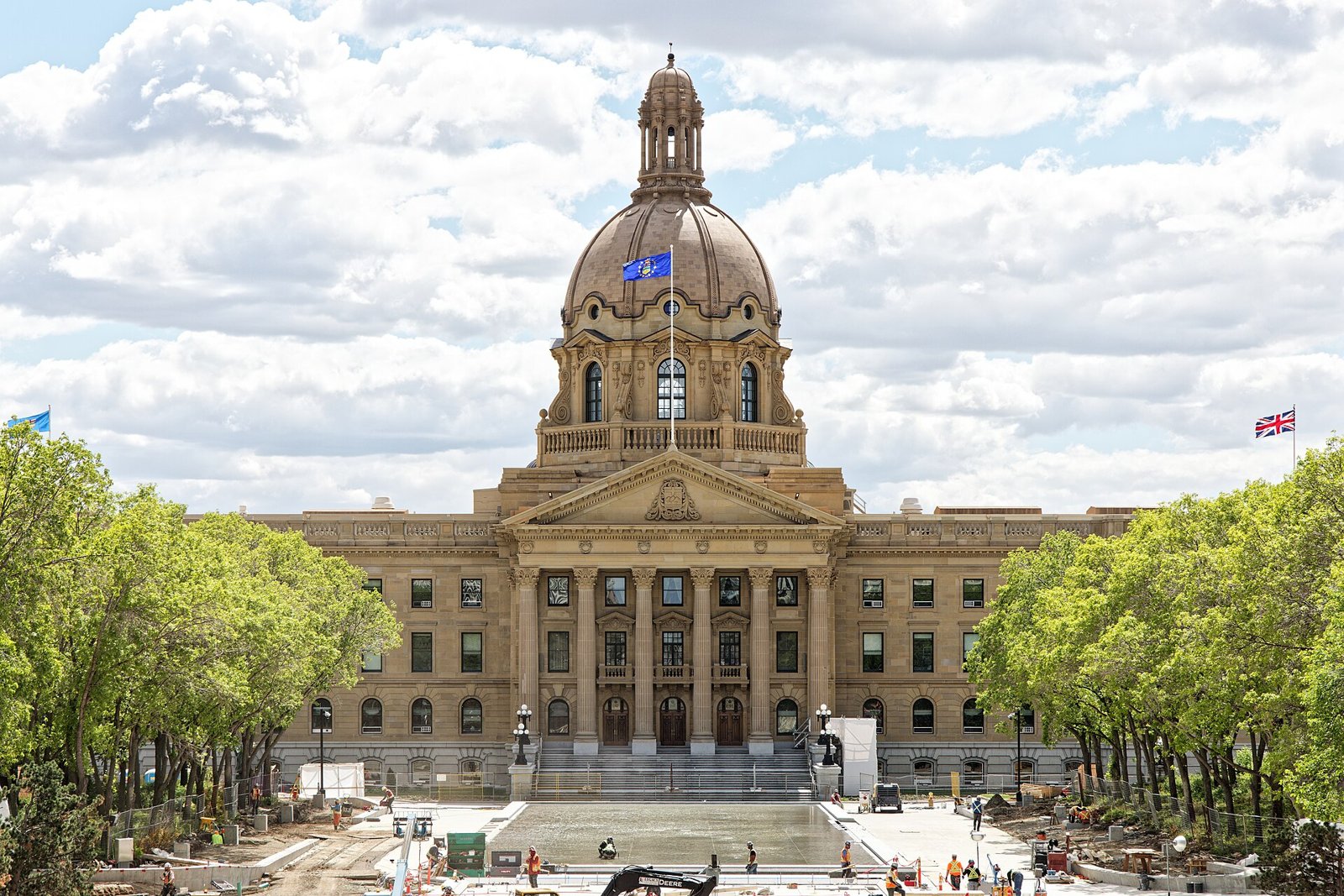 The domed sandstone Alberta Legislature Building in Edmonton under a partly cloudy sky, with trees lining the plaza in front.