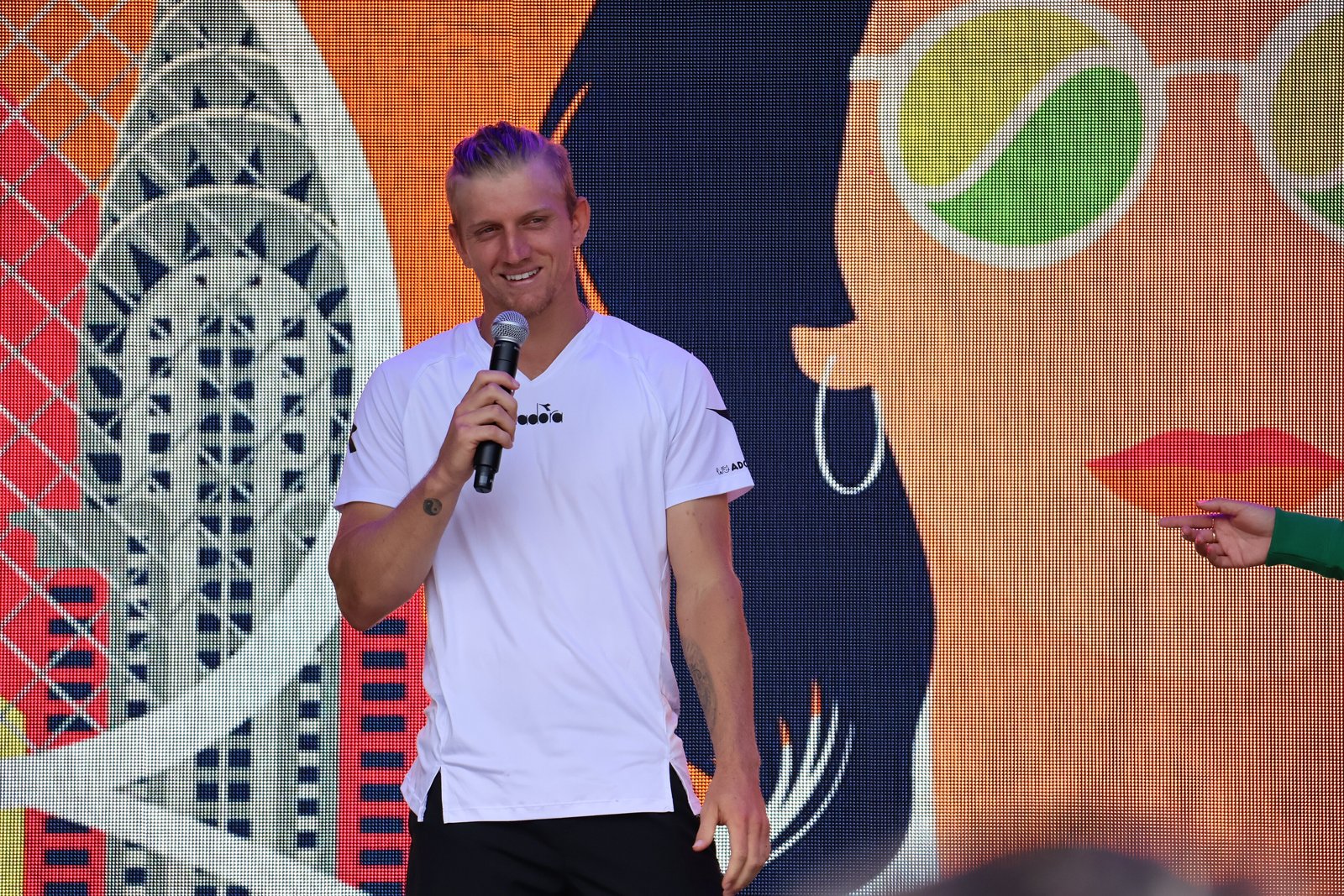 Spanish tennis player Alejandro Davidovich Fokina in a white T-shirt smiling and speaking into a microphone in front of a colorful mural.