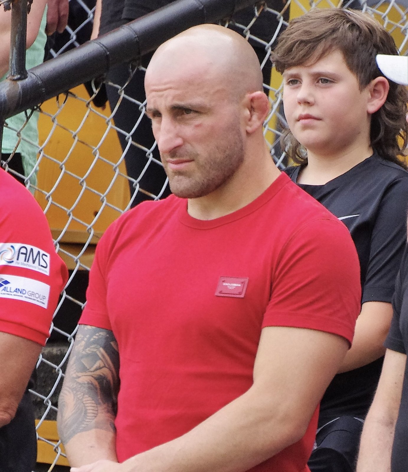 UFC featherweight champion Alexander Volkanovski in a red T-shirt standing beside the cage fence.