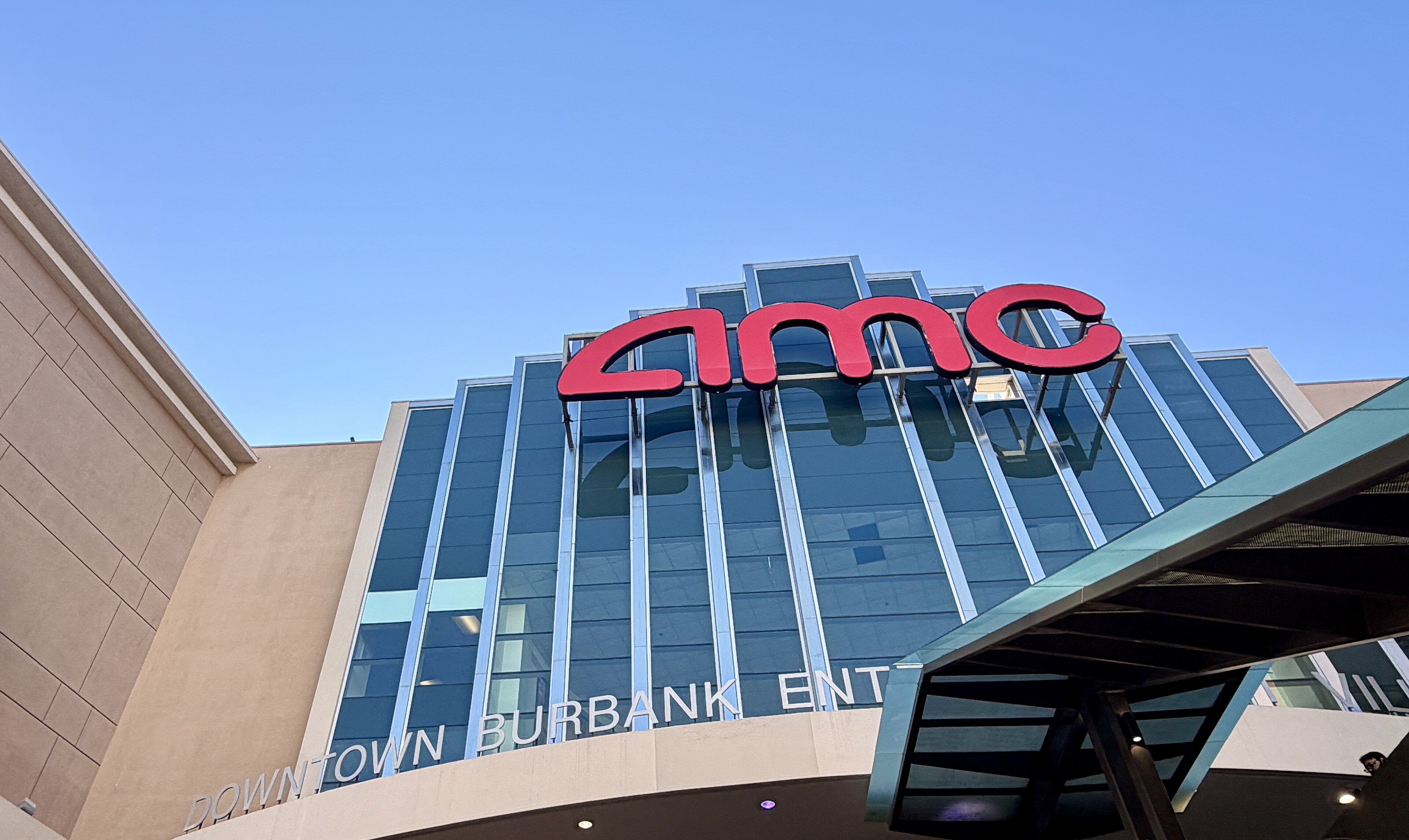 The glass-fronted AMC Burbank 16 multiplex with a large red AMC sign against a clear blue sky, seen from below.