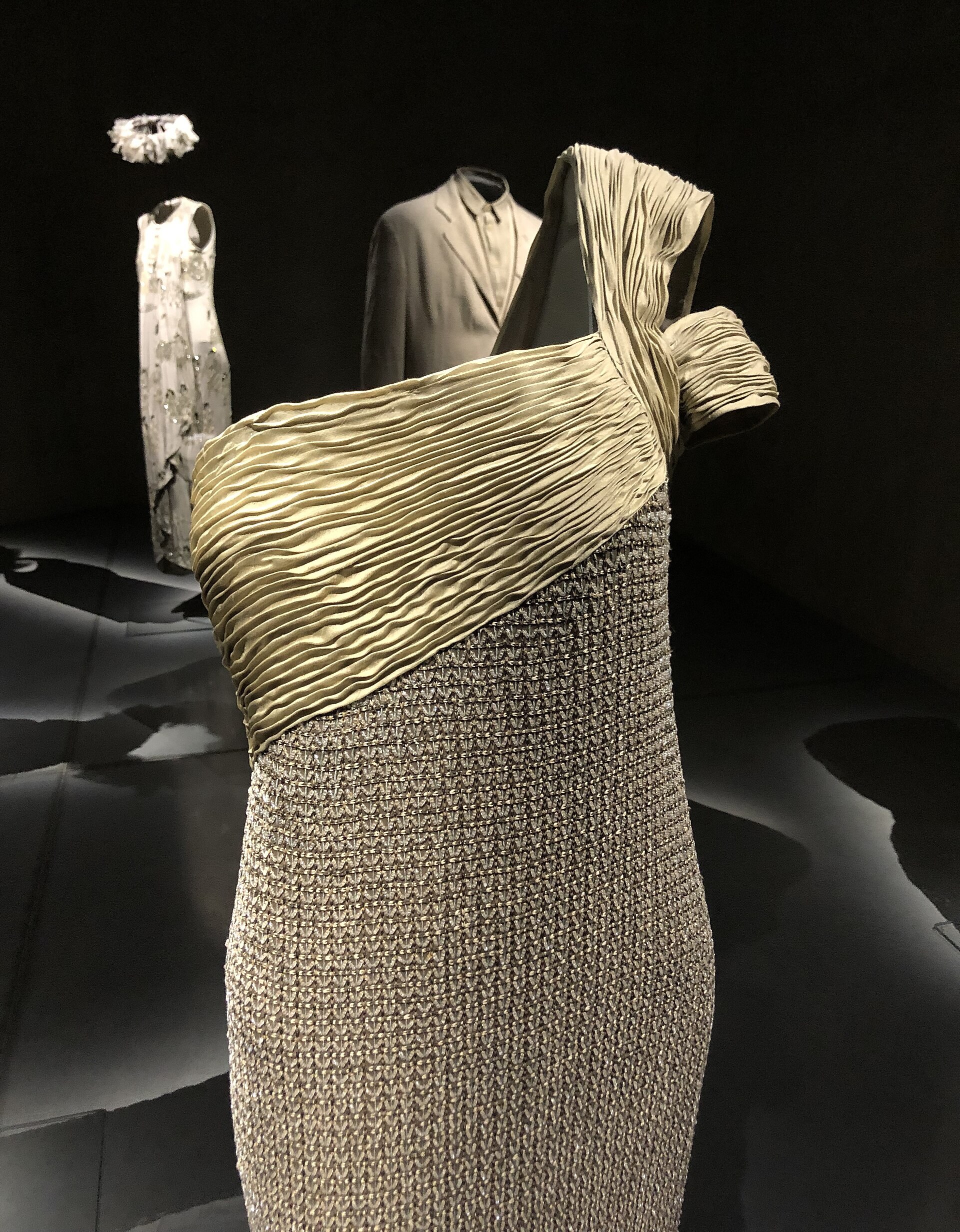 Close-up of a sand-coloured pleated one-shoulder gown with a beaded lower section, displayed on a mannequin against a dark background at the Armani/Silos museum.