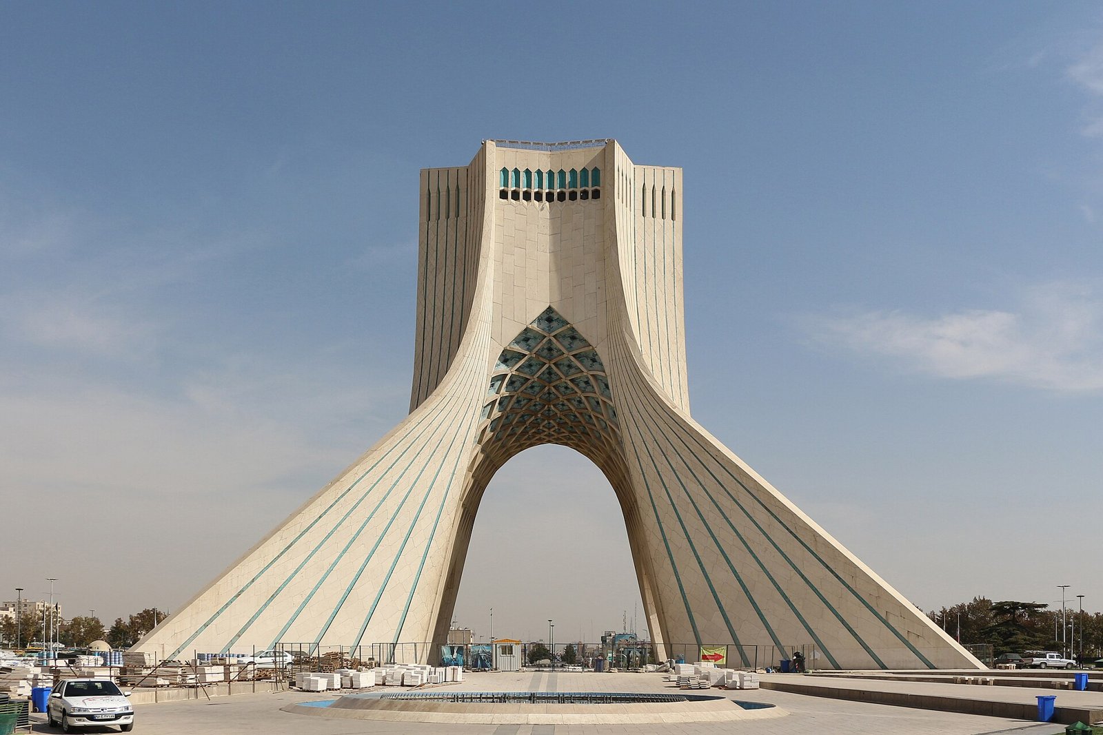 Tehran's white marble Azadi Tower, an arched monument with a wide base and tapering top, under a blue sky with light clouds.