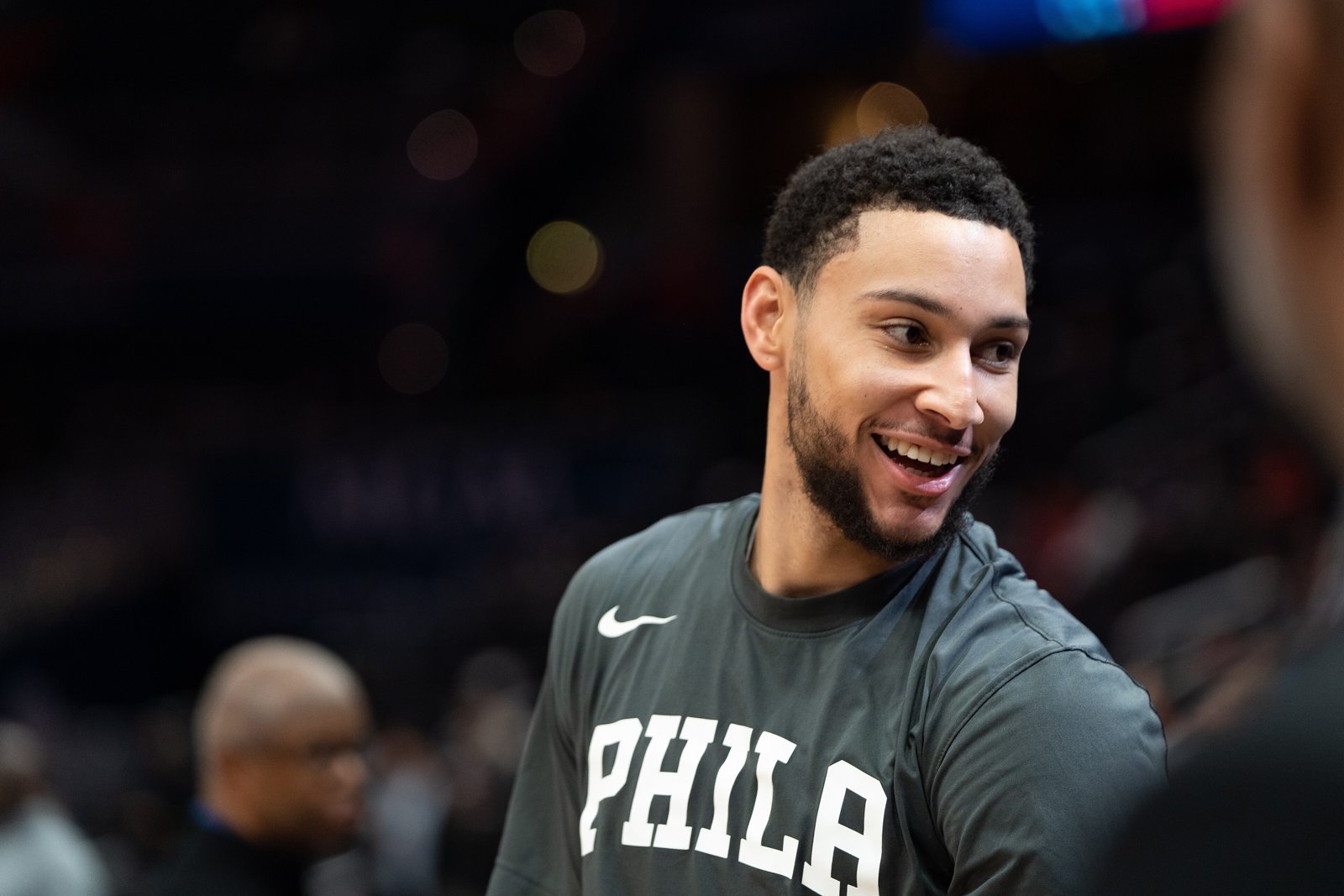 Ben Simmons smiling in a dark Philadelphia warm-up shirt on the court before a game.