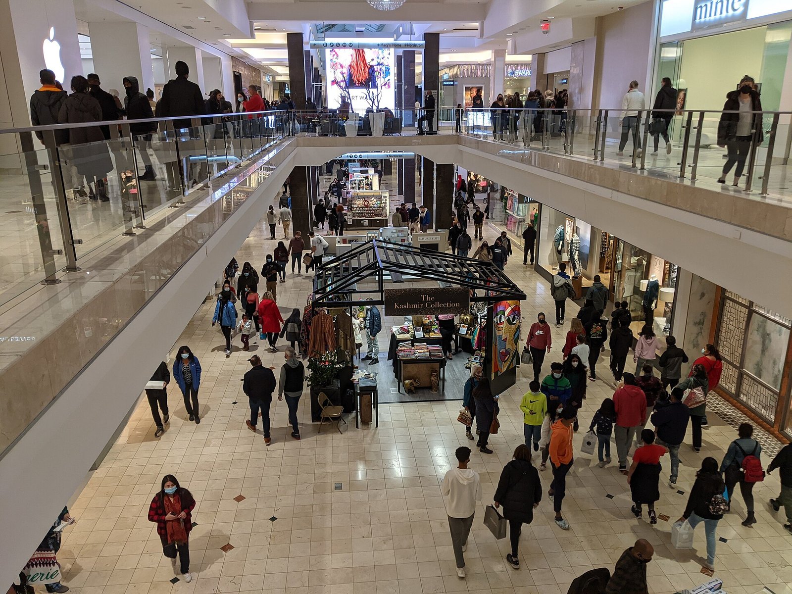 View from an upper floor of a busy two-level shopping mall on Black Friday, with crowds of shoppers walking past kiosks below and store fronts and railings on both sides.