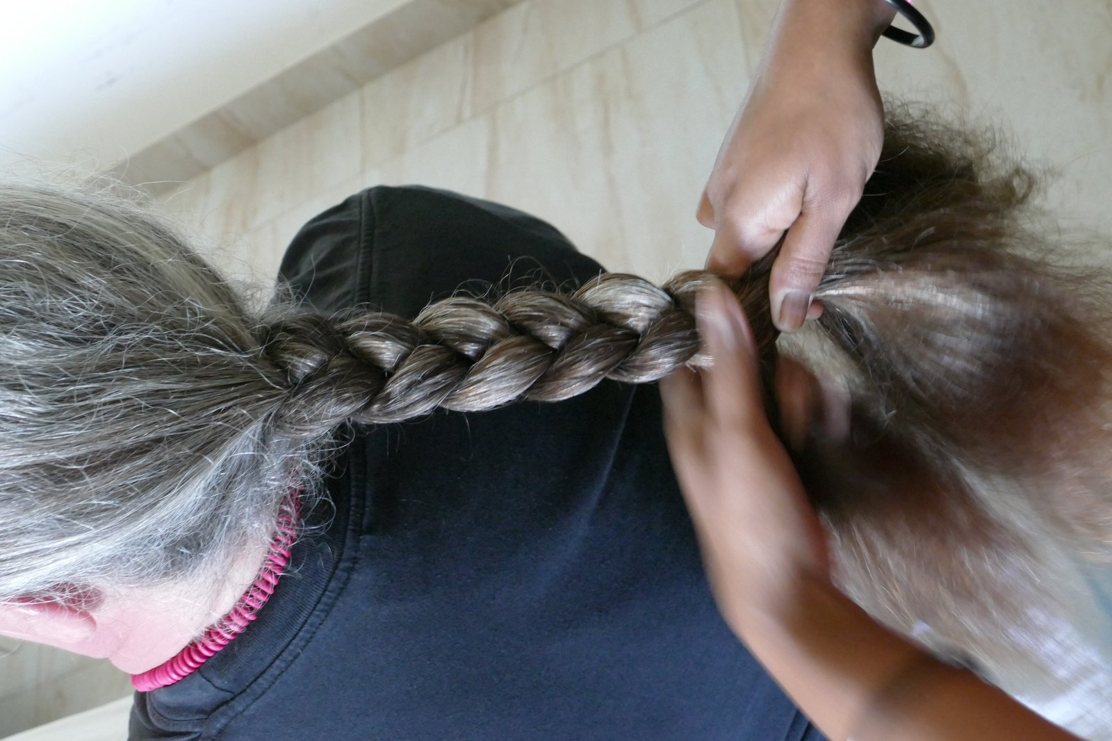 Close-up of hands weaving a long brown-and-grey plait at the back of a person's head.