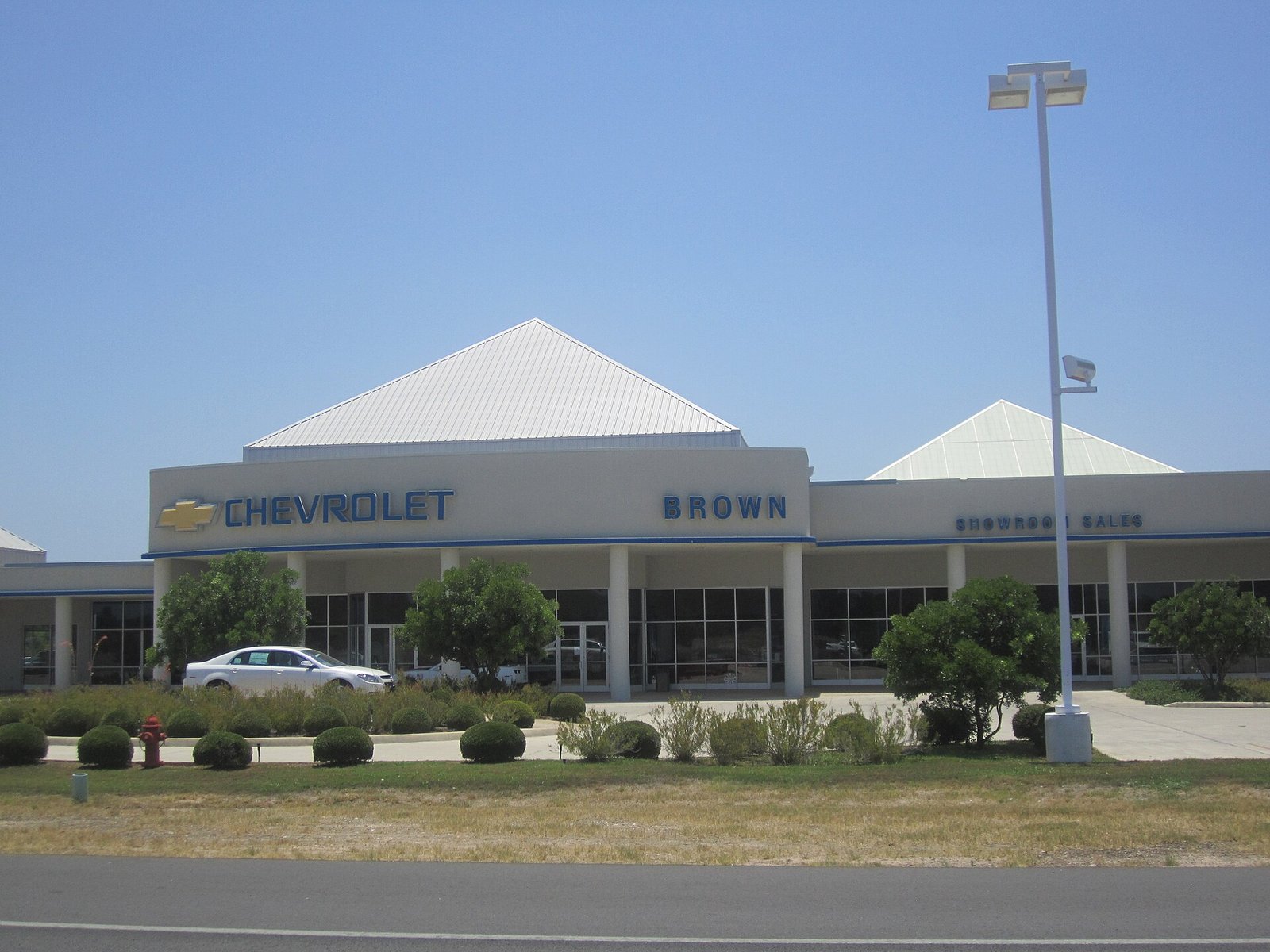 A small-town Chevrolet dealership building with new pickup trucks and cars parked out front under a blue sky.