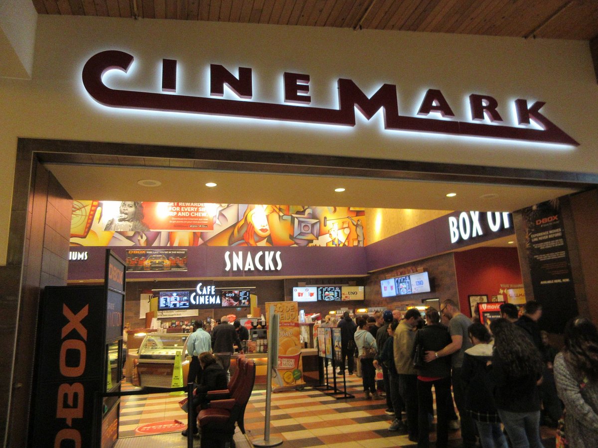 The lobby of a Cinemark theater, with the illuminated Cinemark sign above a snack counter and a line of moviegoers waiting at the concession stand. D-BOX and box office signage is visible on either side.