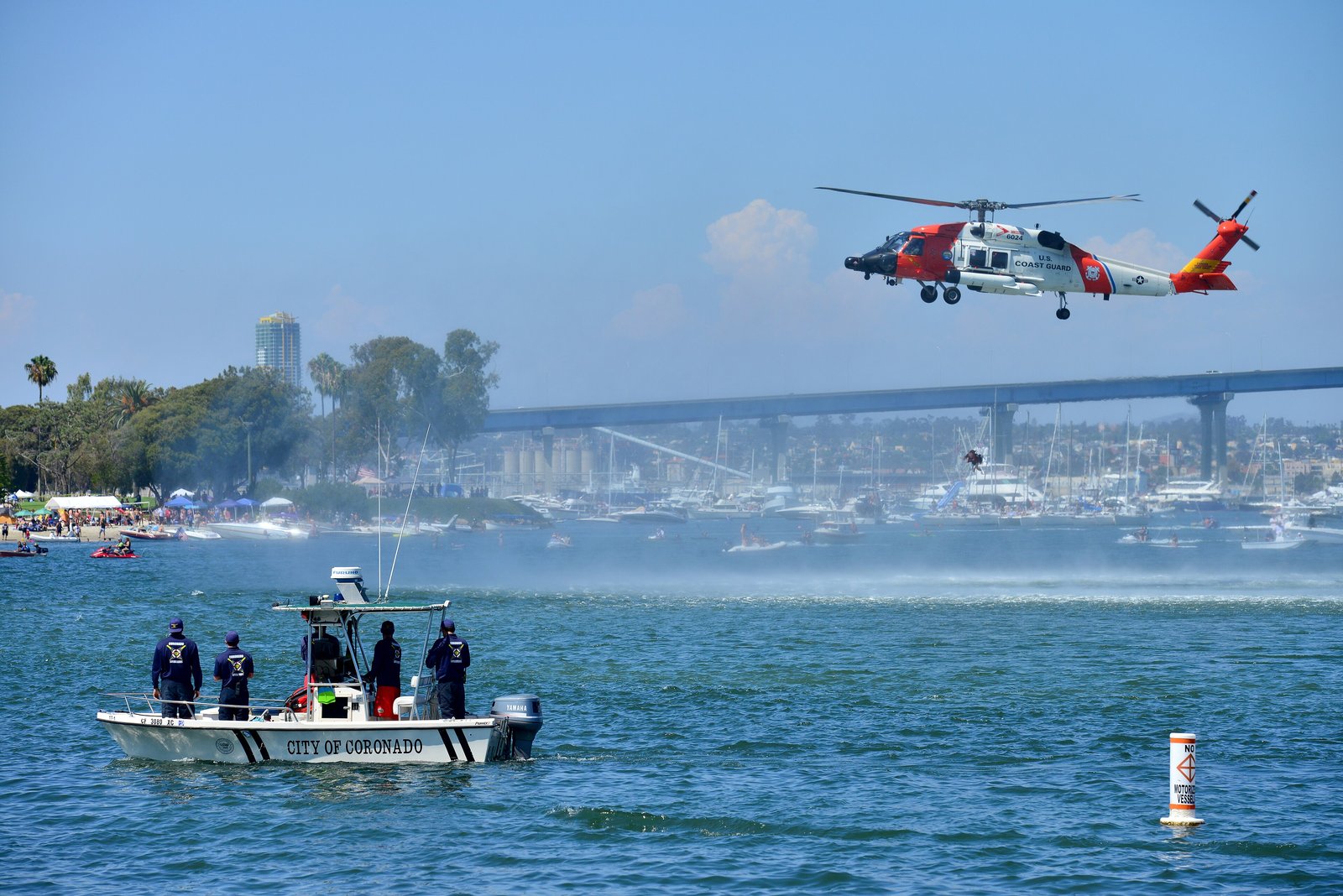 An orange and white U.S. Coast Guard MH-60 Jayhawk helicopter hovering low over water during a rescue demonstration.