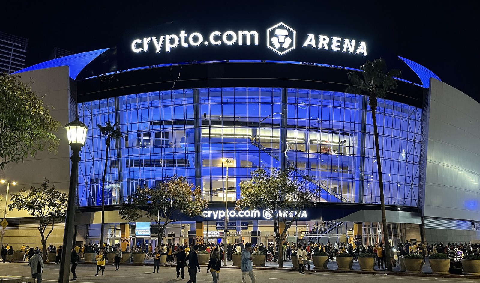 The glass front of Crypto.com Arena lit in blue at night, with its illuminated name across the roof and crowds and palm trees on the plaza outside.