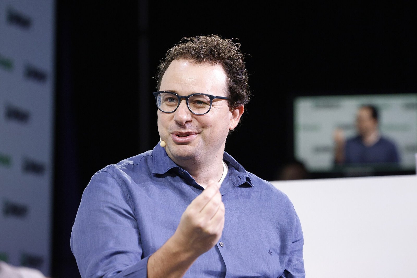 Anthropic CEO Dario Amodei, wearing glasses and a blue shirt, speaking and gesturing on stage at TechCrunch Disrupt 2023.