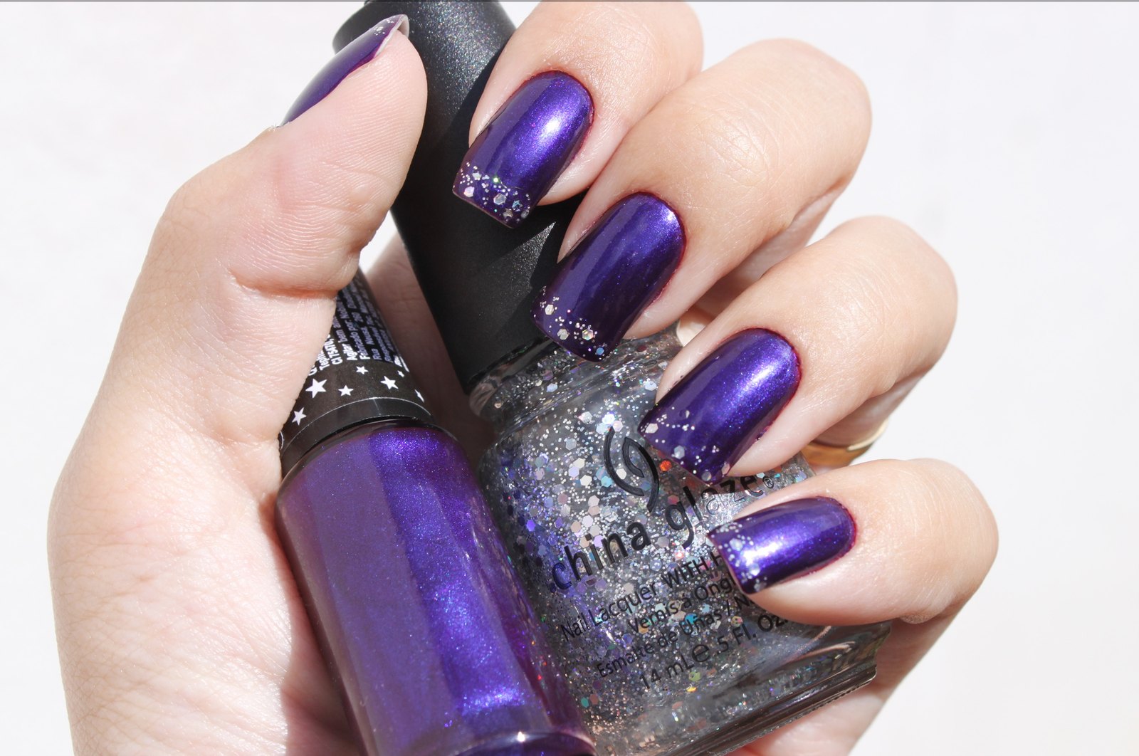 A hand with glossy deep violet nails and silver glitter tips holding two bottles of purple and glitter nail polish against a white background.