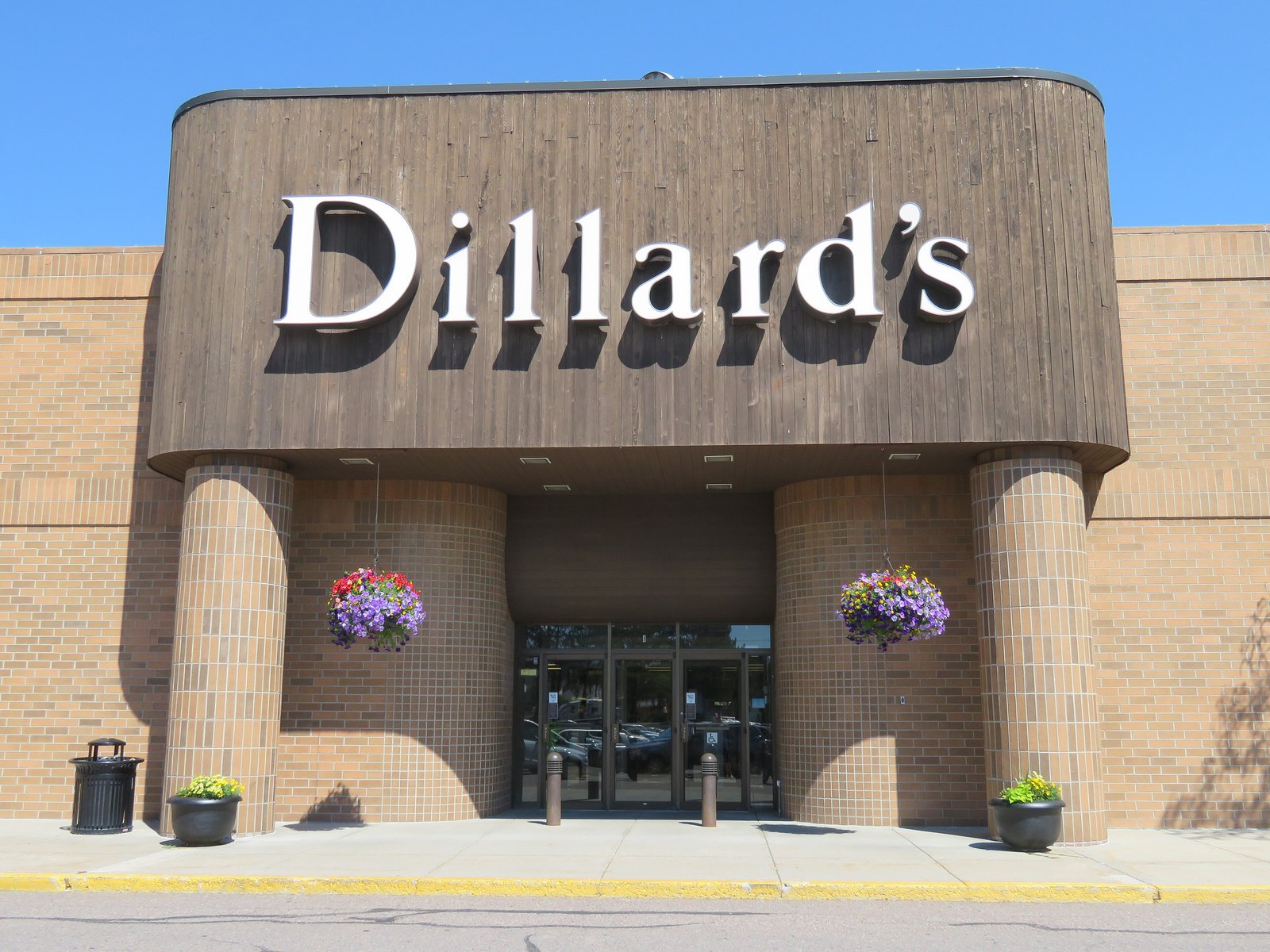The entrance of a Dillard's department store with the brand name in white letters on a brown facade, brick columns and hanging flower baskets.