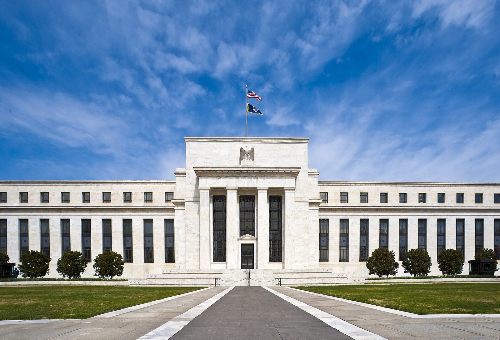 The white marble Eccles Building of the Federal Reserve in Washington under a bright blue sky with wispy clouds, an American flag flying above its columned central entrance and a row of trimmed shrubs in front.