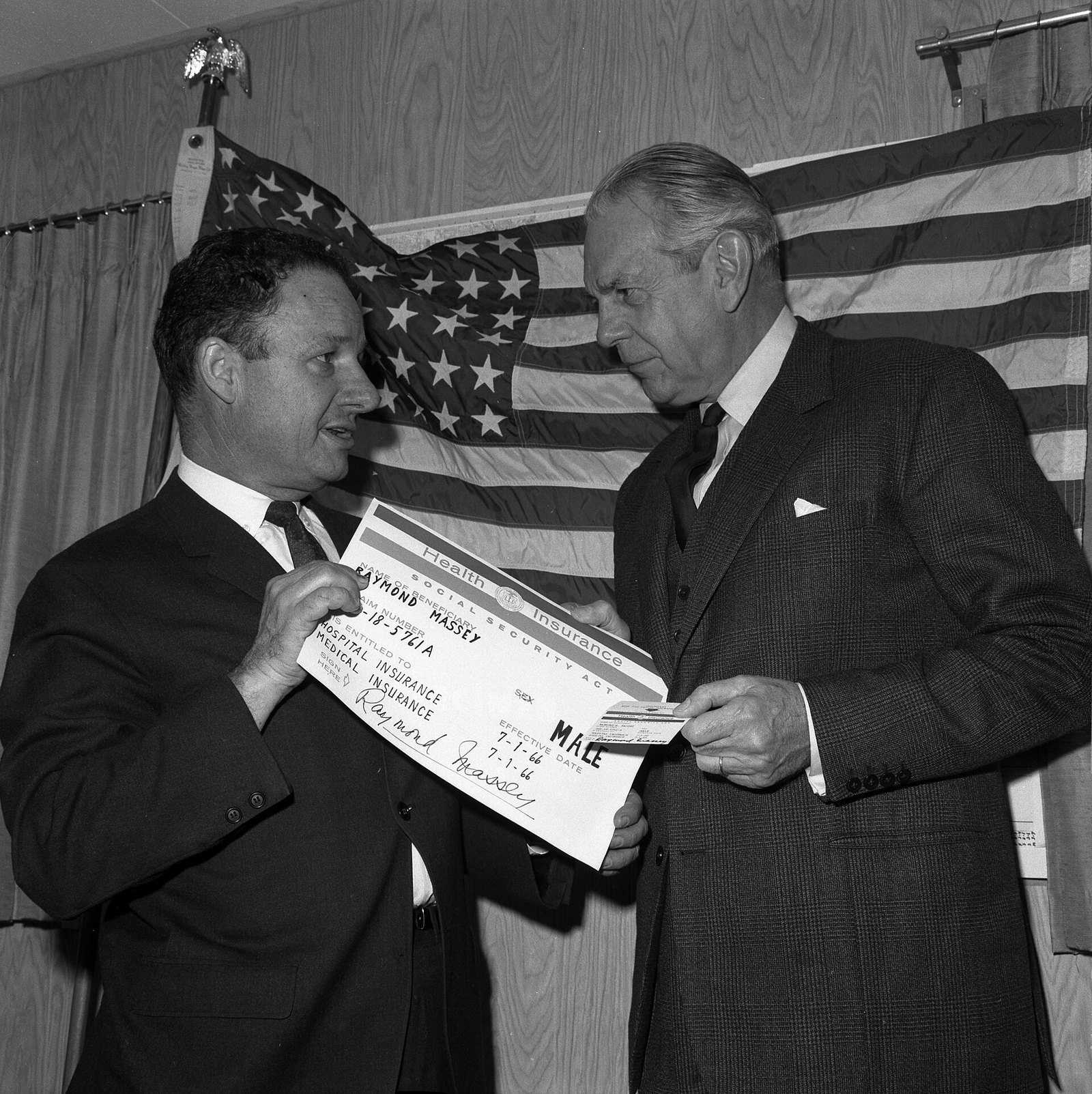 Black-and-white photo of two men in suits in front of an American flag, one holding up an oversized Medicare identification card while the other, older man looks at it.