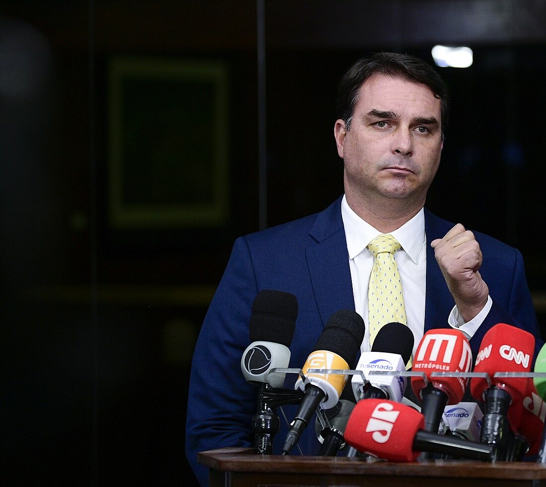 Flávio Bolsonaro, in a navy suit and pale yellow tie, raising a clenched fist as he speaks behind a cluster of broadcast microphones.