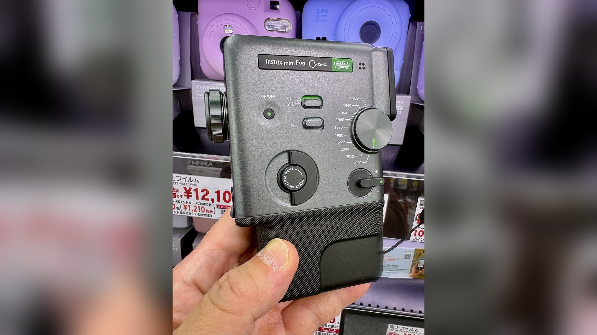 A hand holding the grey Fujifilm Instax mini Evo Cinema camera, showing its side panel with a decade dial labelled 1930 to 2020, a print dial and a still/cine switch, in front of a shelf of pastel Instax cameras.
