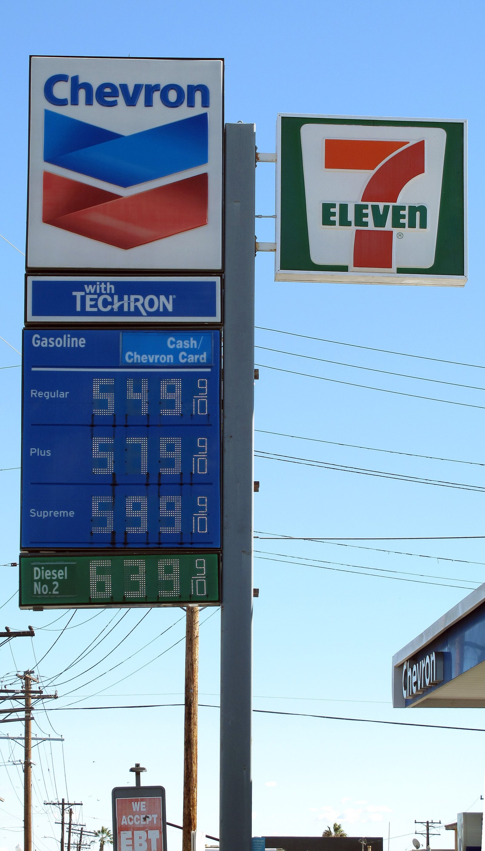 A tall Chevron gas station sign against a blue sky listing gasoline prices of $5.49, $5.79 and $5.99 a gallon and diesel at $6.39, next to a 7-Eleven sign.