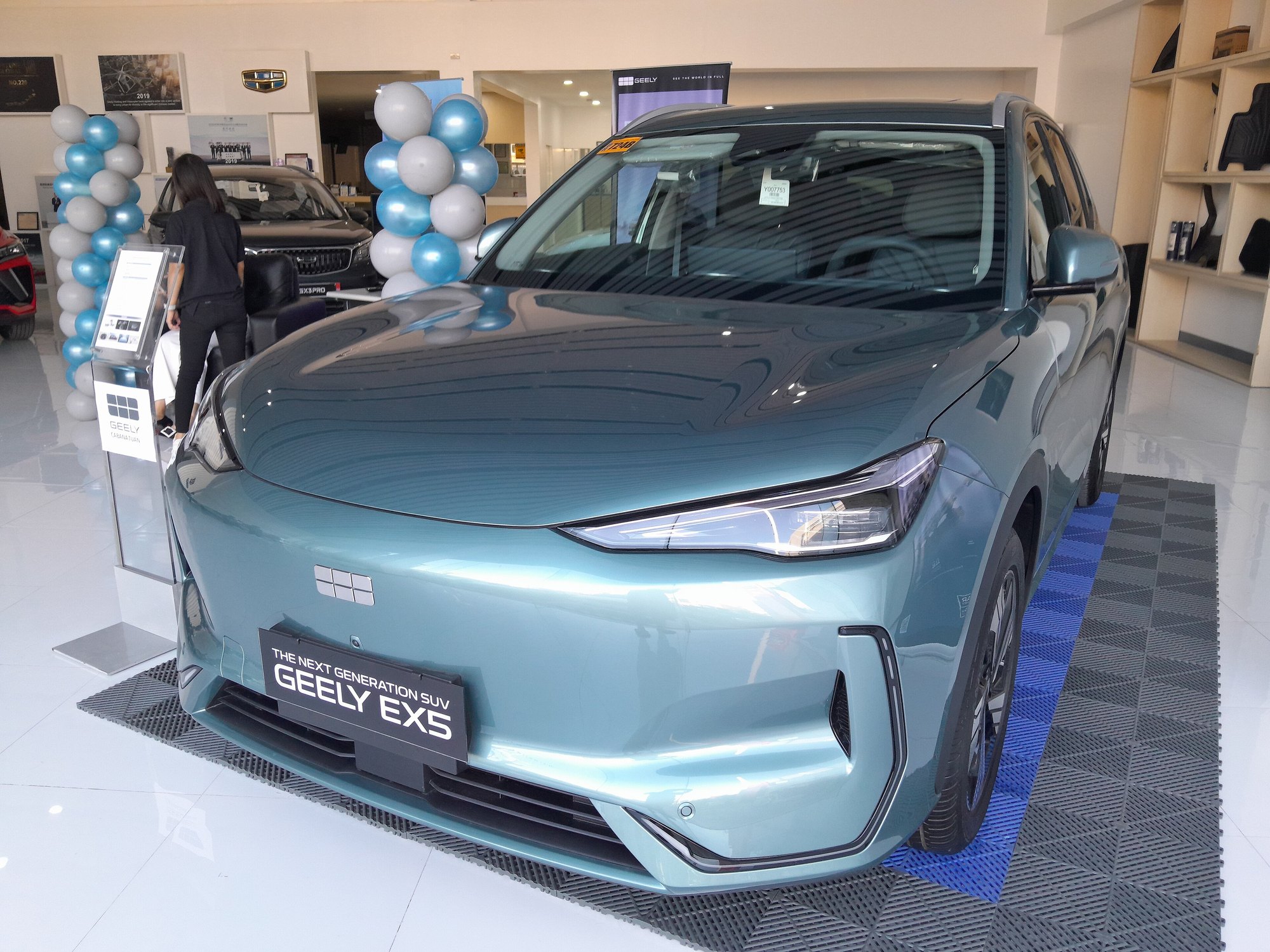 A teal Geely EX5 electric SUV on a dealership showroom floor, with a sign reading The Next Generation SUV Geely EX5 and blue and white balloons behind it.