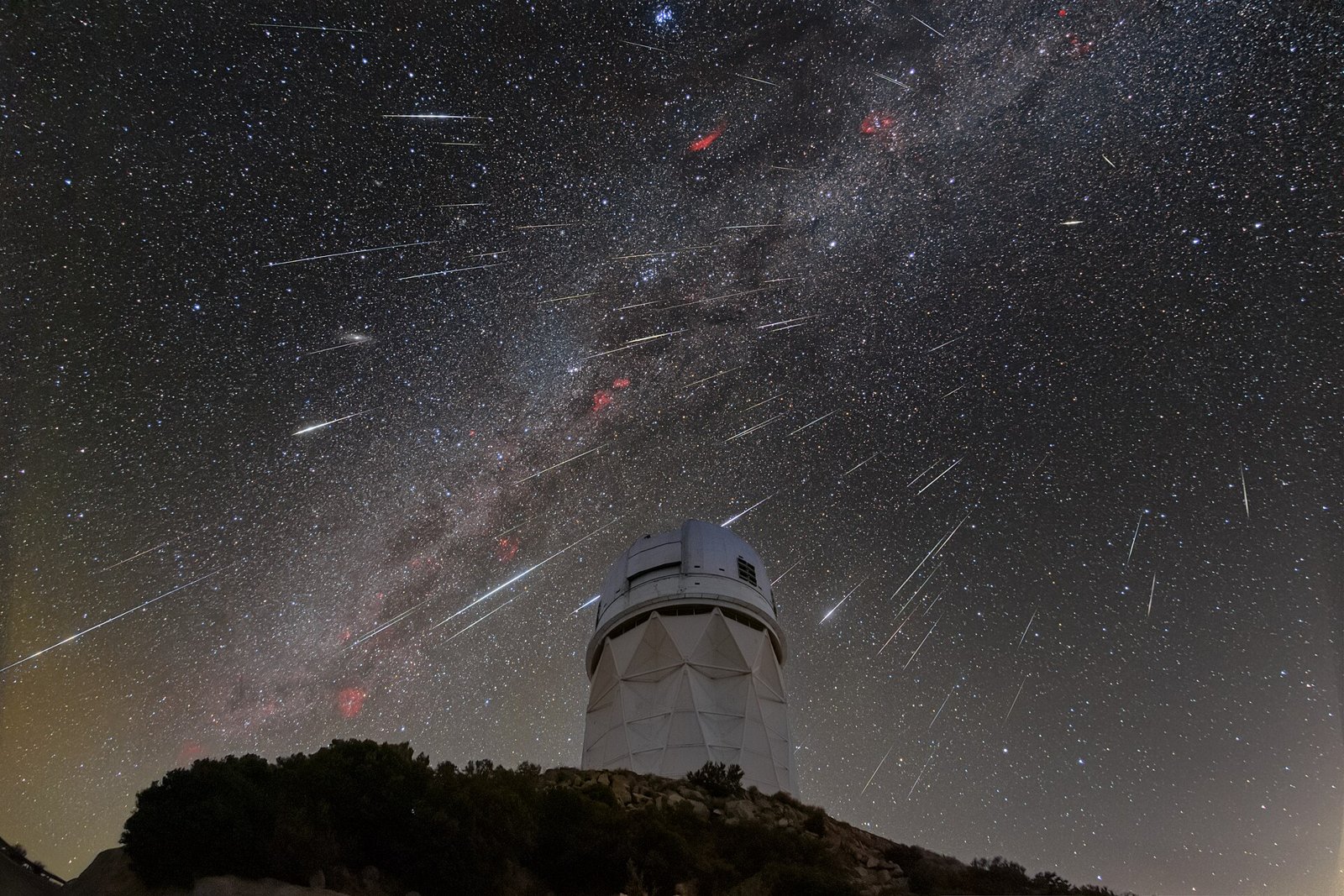 Dozens of Geminid meteor streaks radiating across a starry sky and the Milky Way above a white observatory dome on a dark ridge.