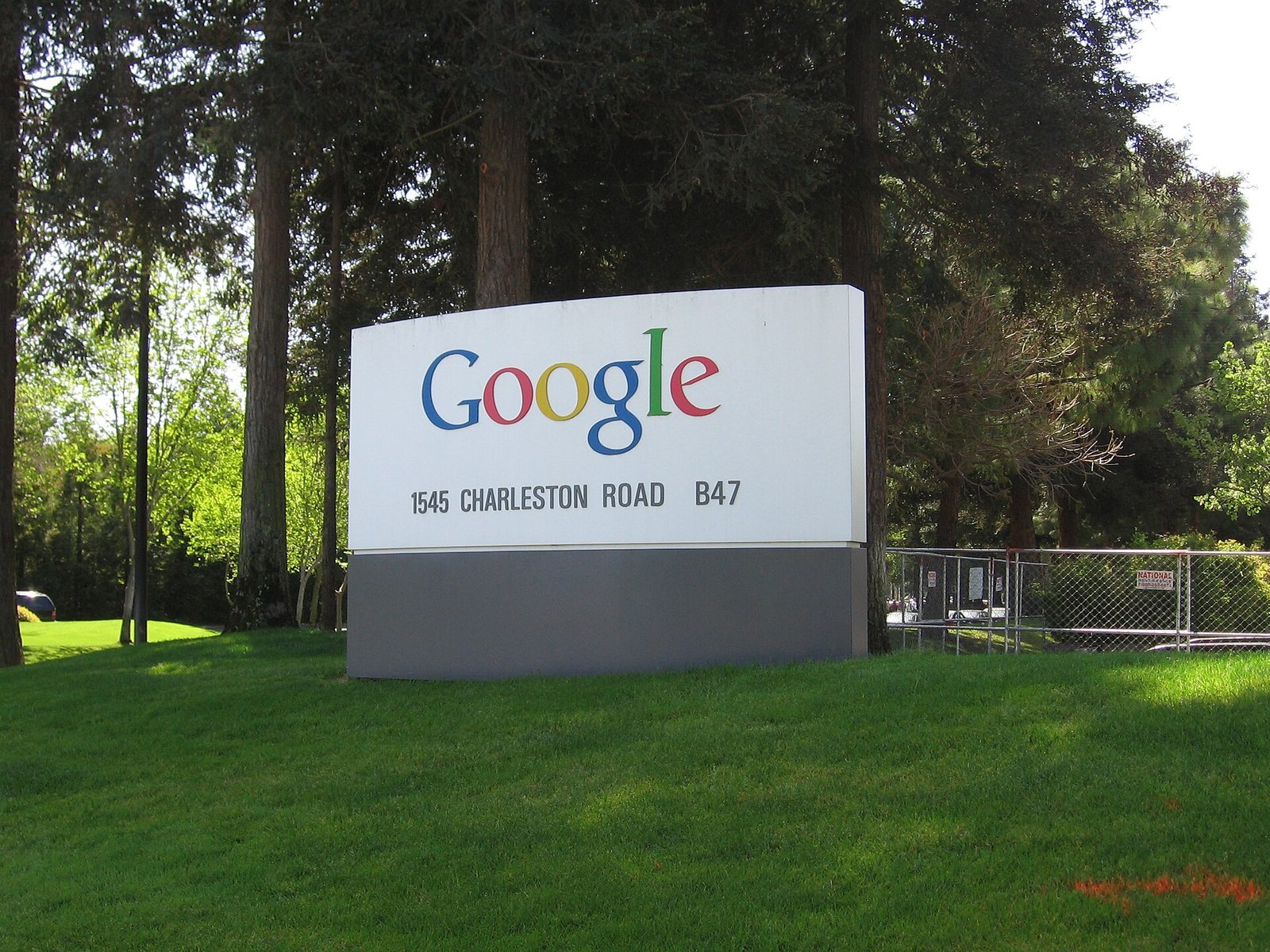 A white Google sign with the colorful Google logo and the address 1545 Charleston Road on a lawn in front of trees at the Googleplex.