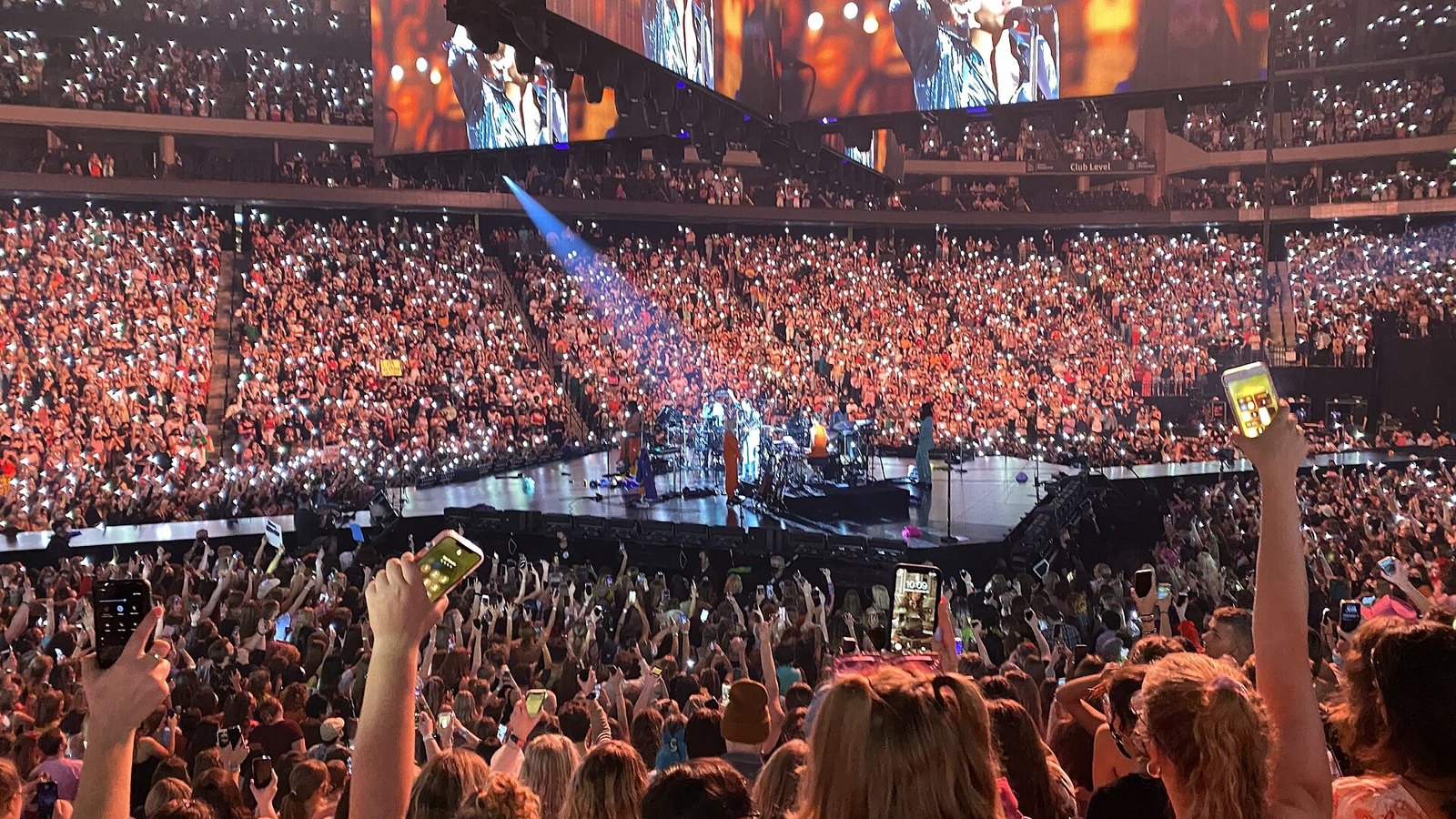 A packed arena glows pink and gold with phone lights as Harry Styles performs on a distant stage beneath large video screens.