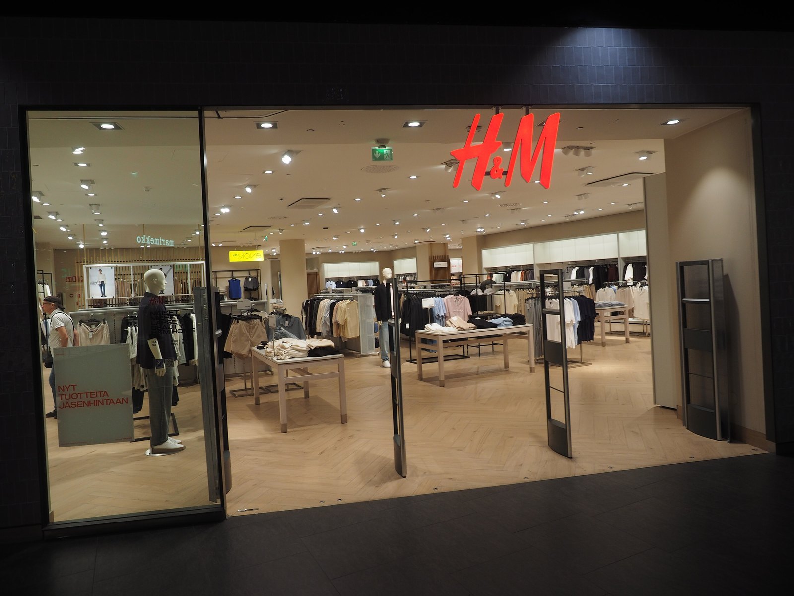 The glass entrance of an H&M store inside a shopping centre, with the red H&M logo above and racks of clothing and mannequins visible inside.