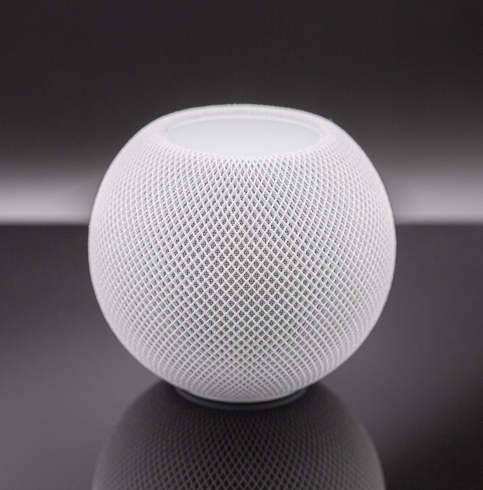 Close-up of a round white Apple HomePod mini smart speaker with its mesh fabric cover against a dark gray background.