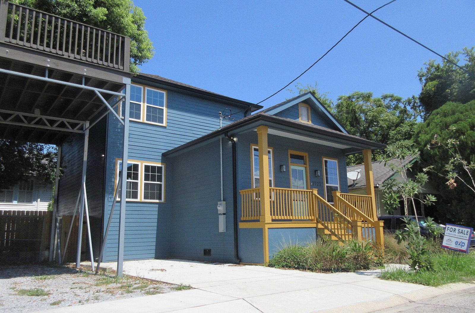 A new two-storey blue house with a yellow-trimmed front porch and steps, a small for-sale sign in the front yard and trees behind it under a clear blue sky.