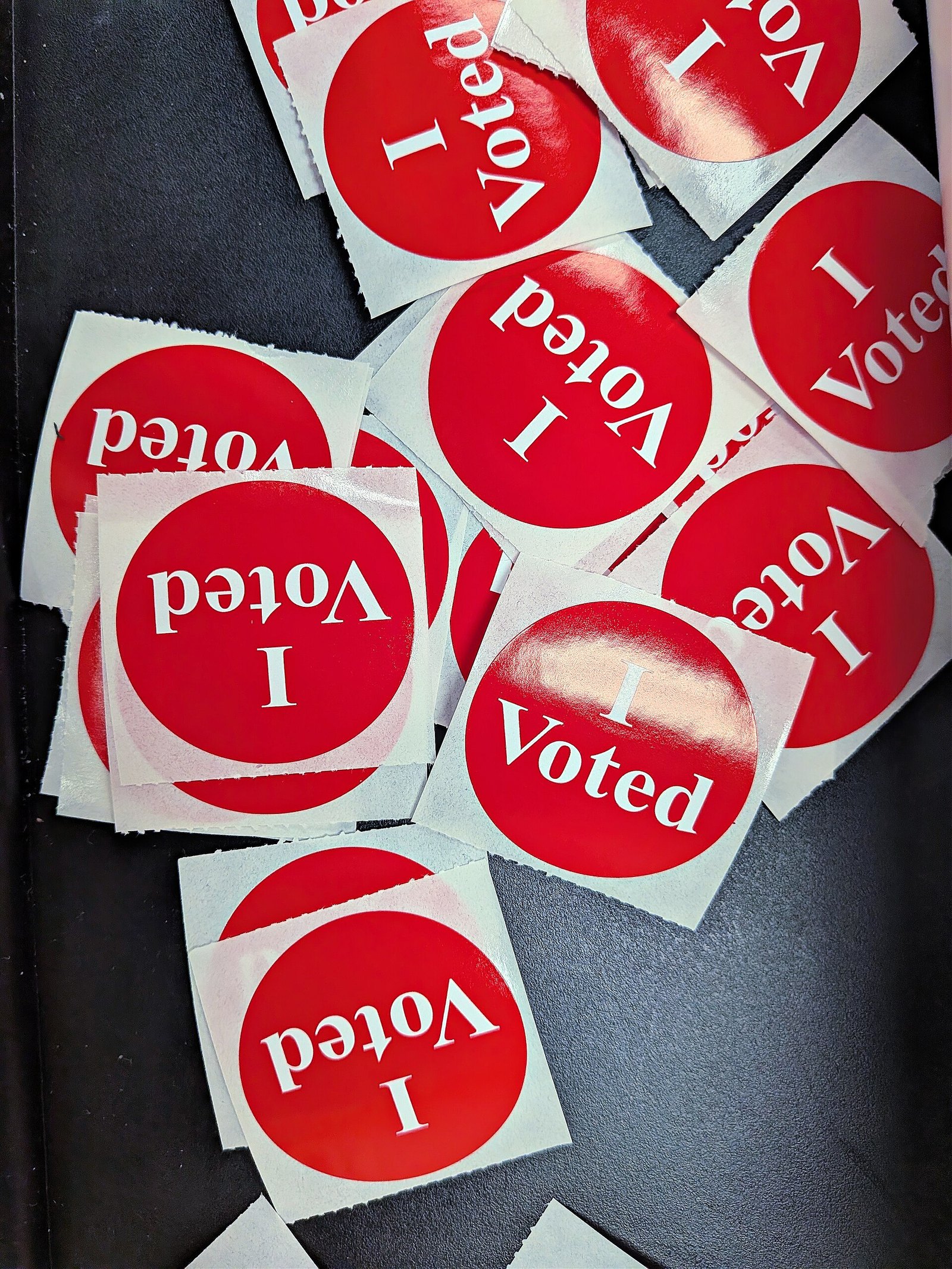 A scattered pile of round red and white I Voted stickers on a dark surface at a polling station.