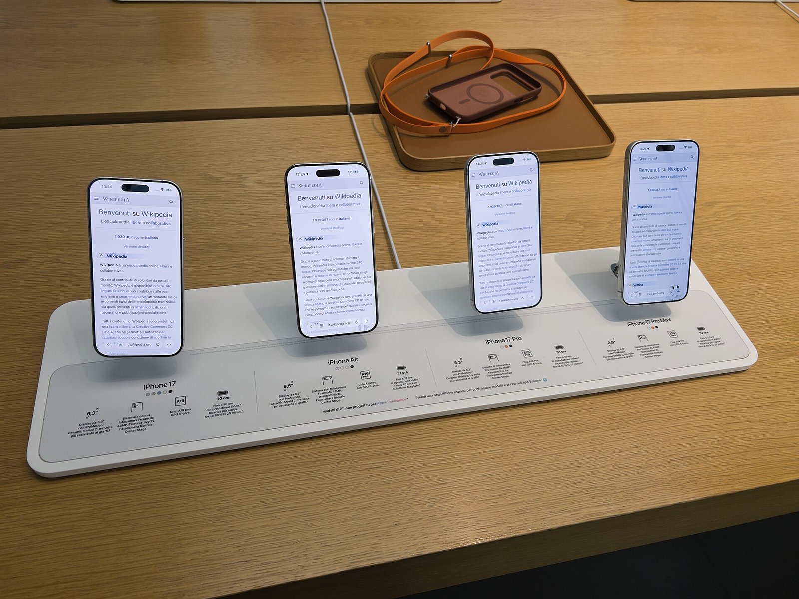 Four iPhones from the iPhone 17 lineup standing upright on a white display stand on a wooden Apple Store table.