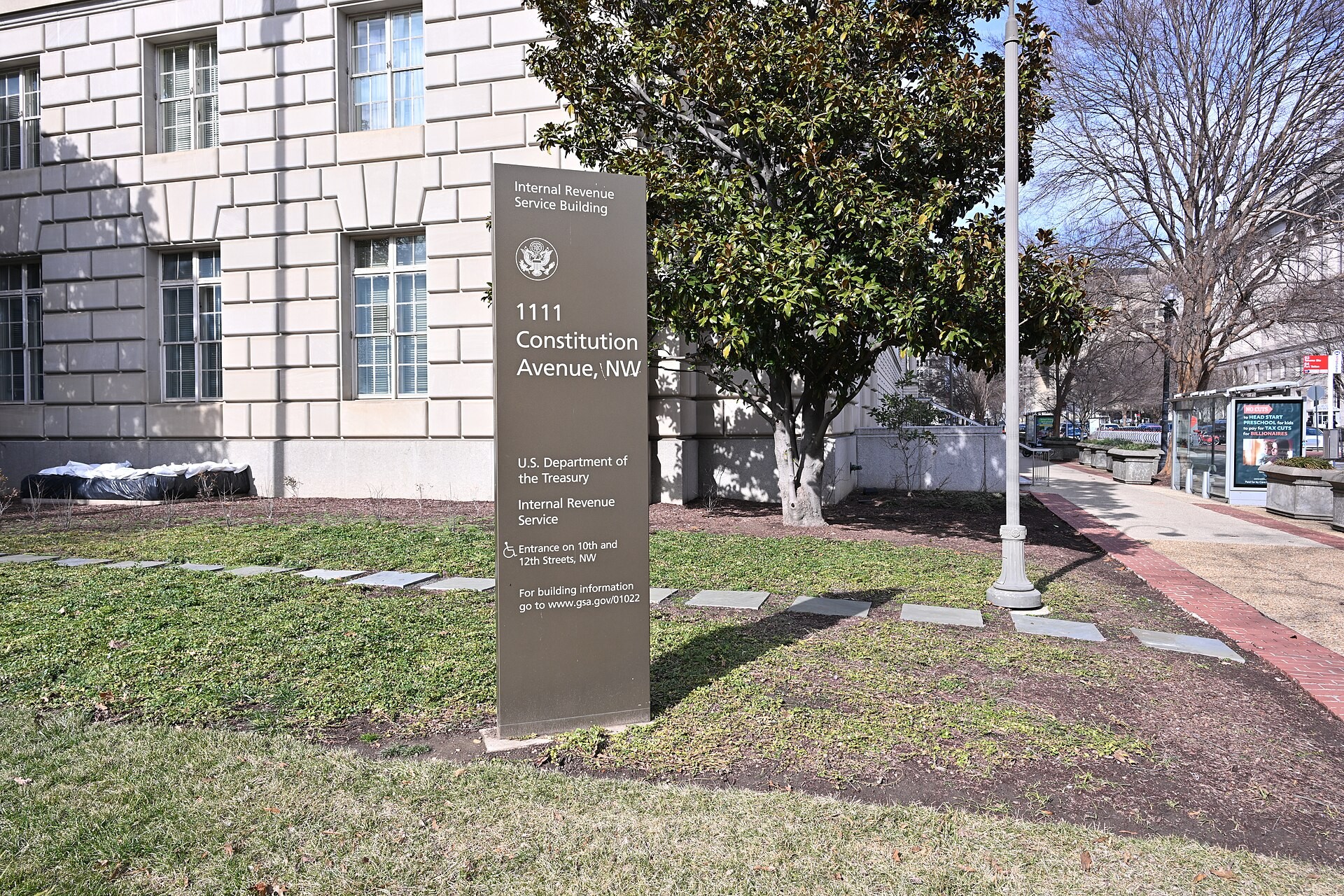 A tall brown sign on a lawn reading Internal Revenue Service Building, 1111 Constitution Avenue NW, U.S. Department of the Treasury, Internal Revenue Service, in front of a pale stone government building and a magnolia tree.