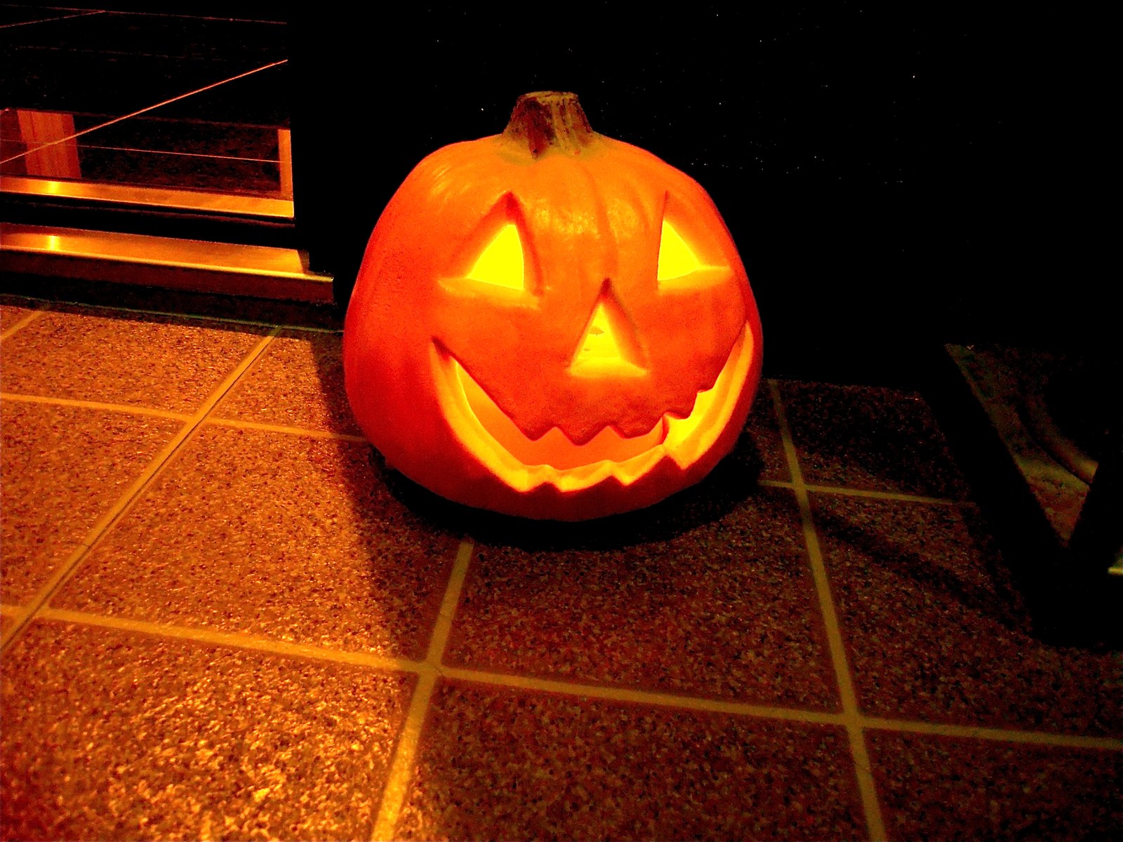 A glowing carved pumpkin with triangle eyes and a toothy smile sitting on a tiled step outside a doorway at night.