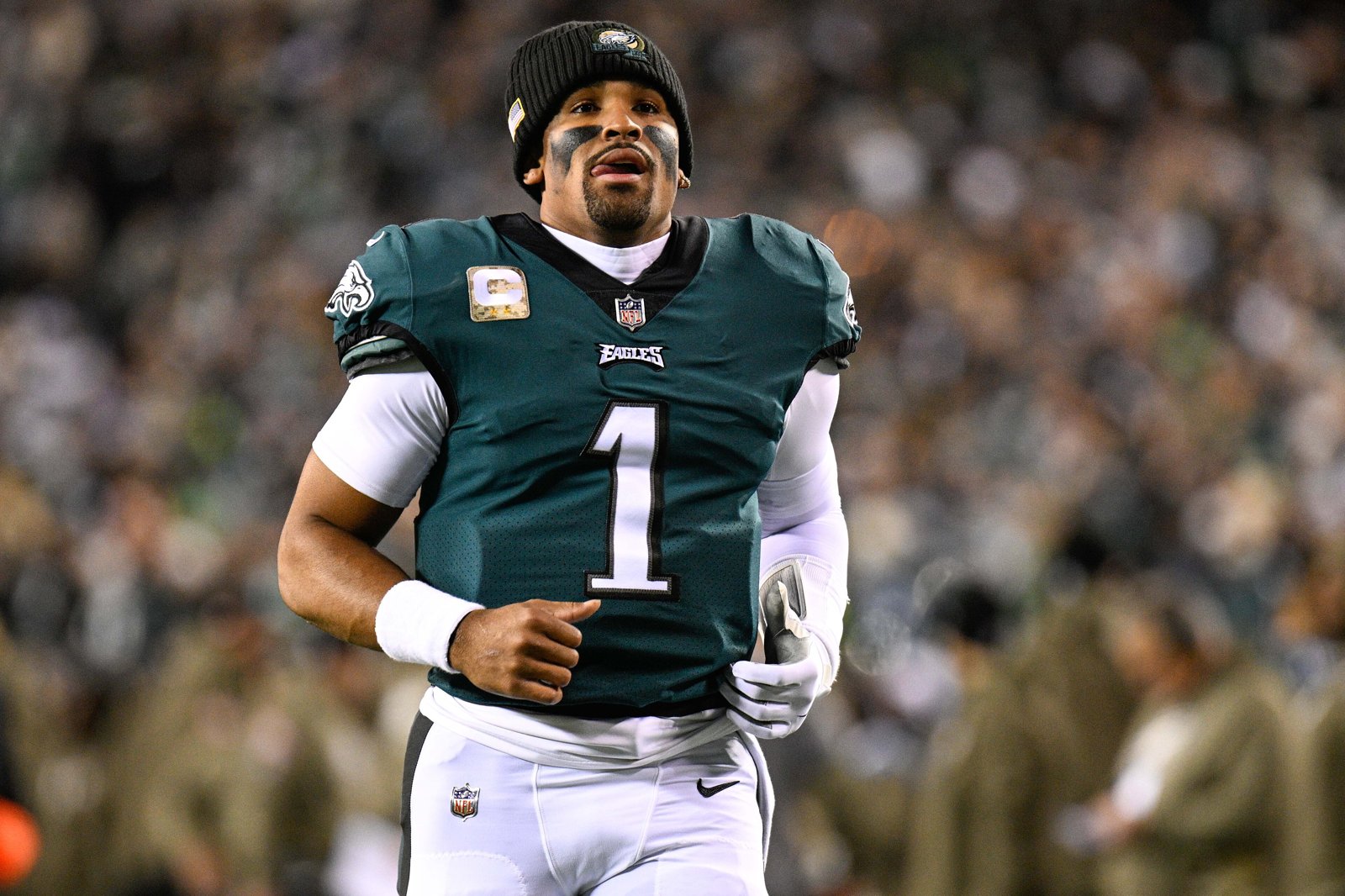 Philadelphia Eagles quarterback Jalen Hurts in a green No. 1 jersey and black beanie jogging on the field in front of a crowd.