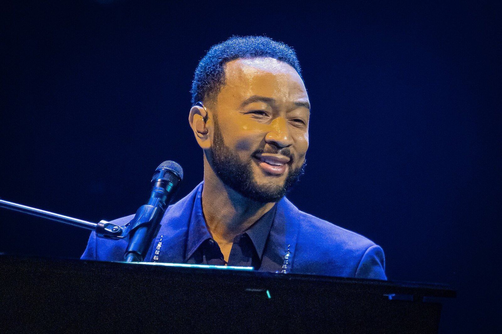 John Legend smiles while seated at a piano behind a microphone, wearing a dark blue jacket under deep blue stage lighting.
