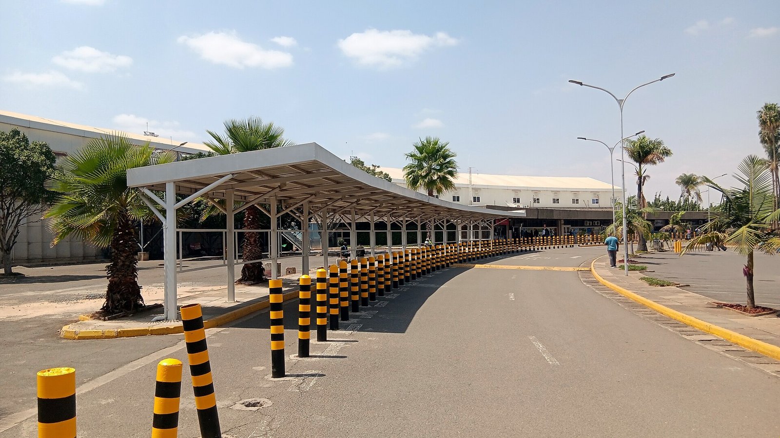 The landside approach to the terminal at Jomo Kenyatta International Airport in Nairobi, with a covered walkway, palm trees and yellow-and-black bollards.