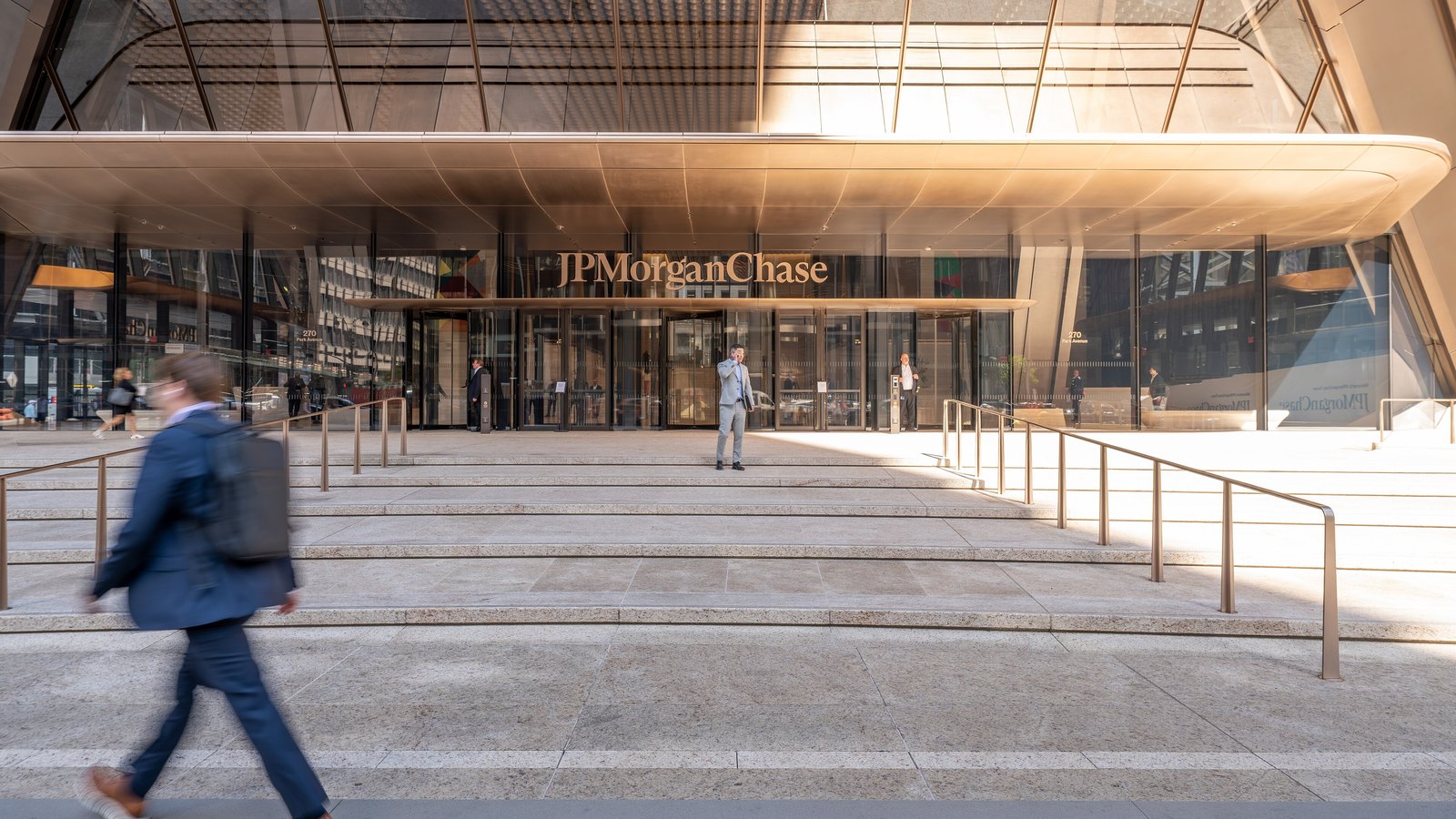 A man in a suit walks past wide stone steps leading to the glass entrance of JPMorgan Chase's headquarters at 270 Park Avenue, under a curved metal canopy with the JPMorganChase name above the doors.