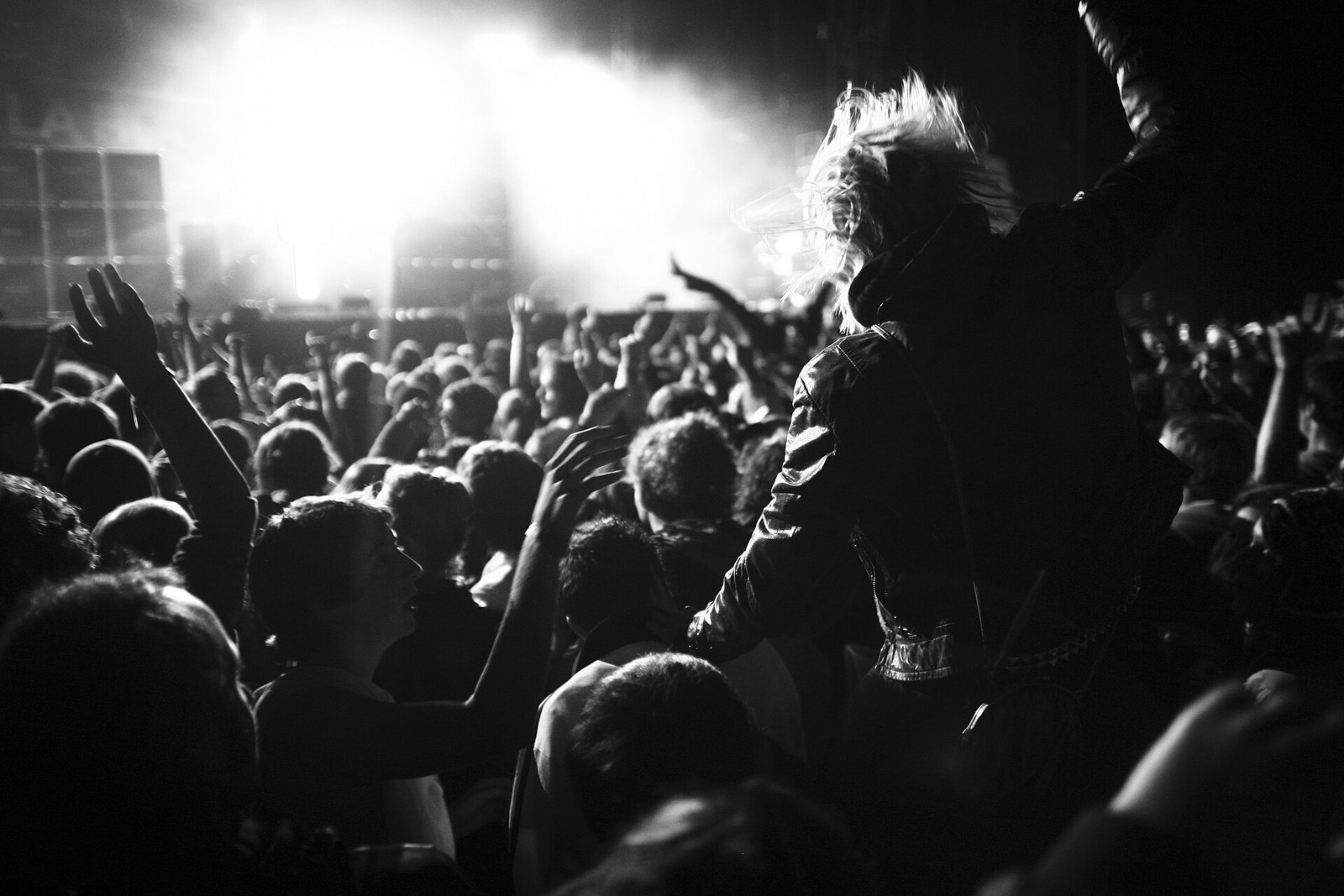 Black-and-white photo of a packed crowd with raised arms at a Justice concert, a bright stage behind them and a long-haired fan in the foreground.