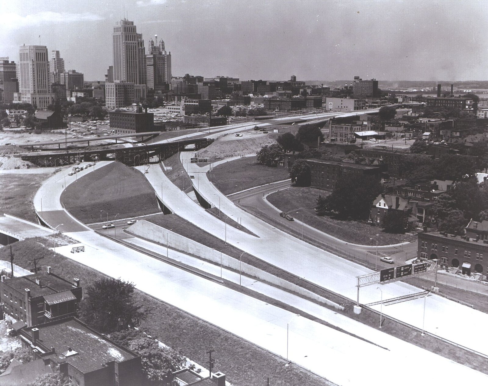 Black-and-white aerial view of curving interstate ramps and overpasses leading into downtown Kansas City, with office towers in the distance.