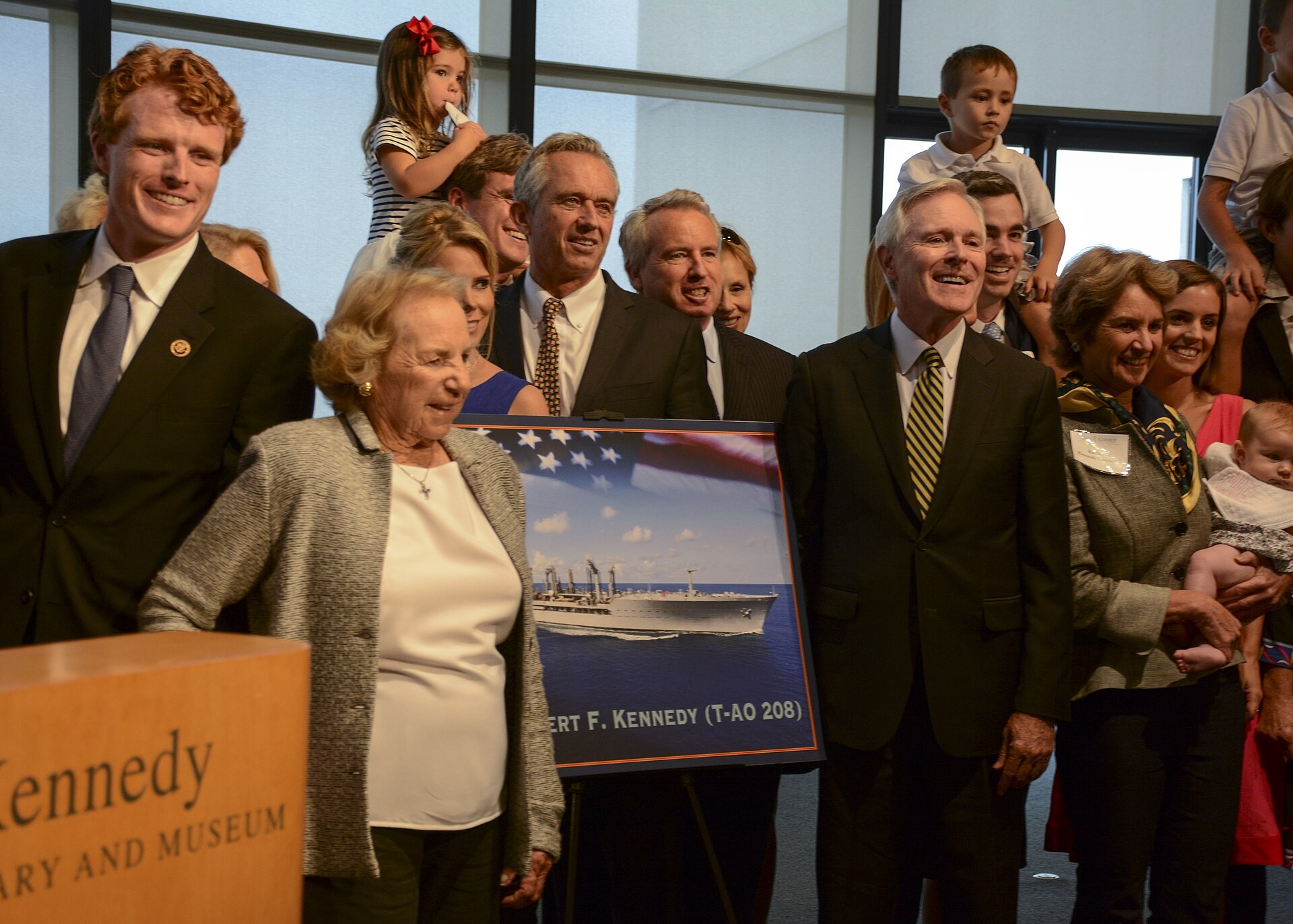 Ethel Kennedy, an elderly woman in a gray jacket, stands with smiling Kennedy family members and Navy Secretary Ray Mabus beside a framed picture of the ship USNS Robert F. Kennedy