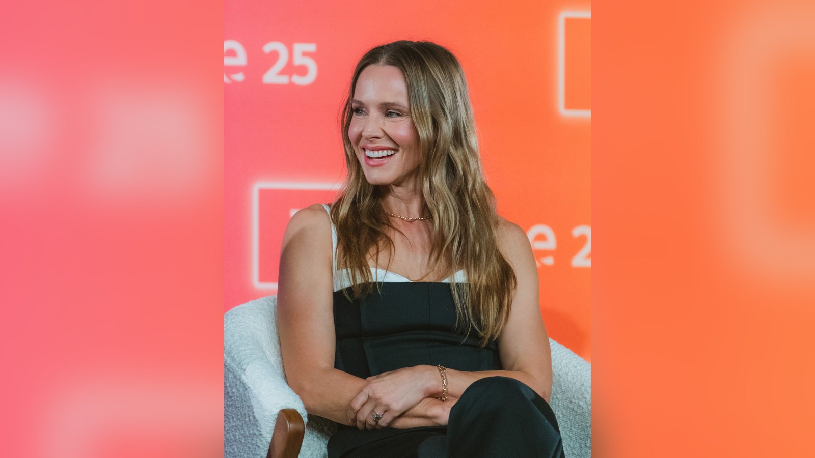 Kristen Bell, with long wavy blonde hair and a black-and-white top, smiles while seated on stage during a Nobody Wants This panel.