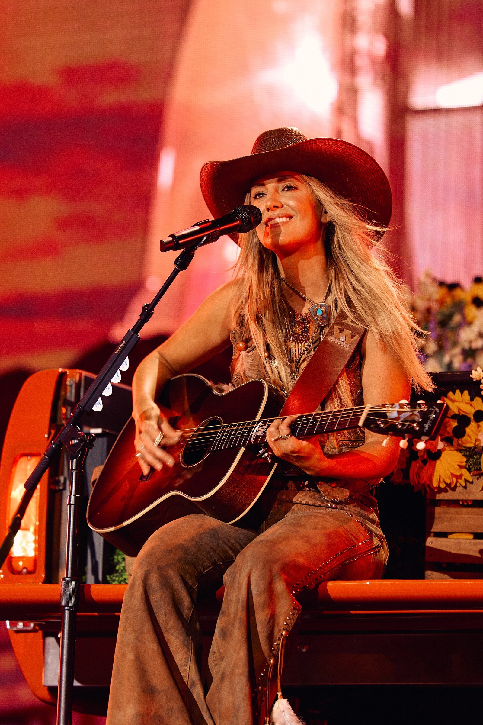 Lainey Wilson in a wide-brimmed cowboy hat and flared trousers sits on stage playing an acoustic guitar and singing into a microphone under warm red light.