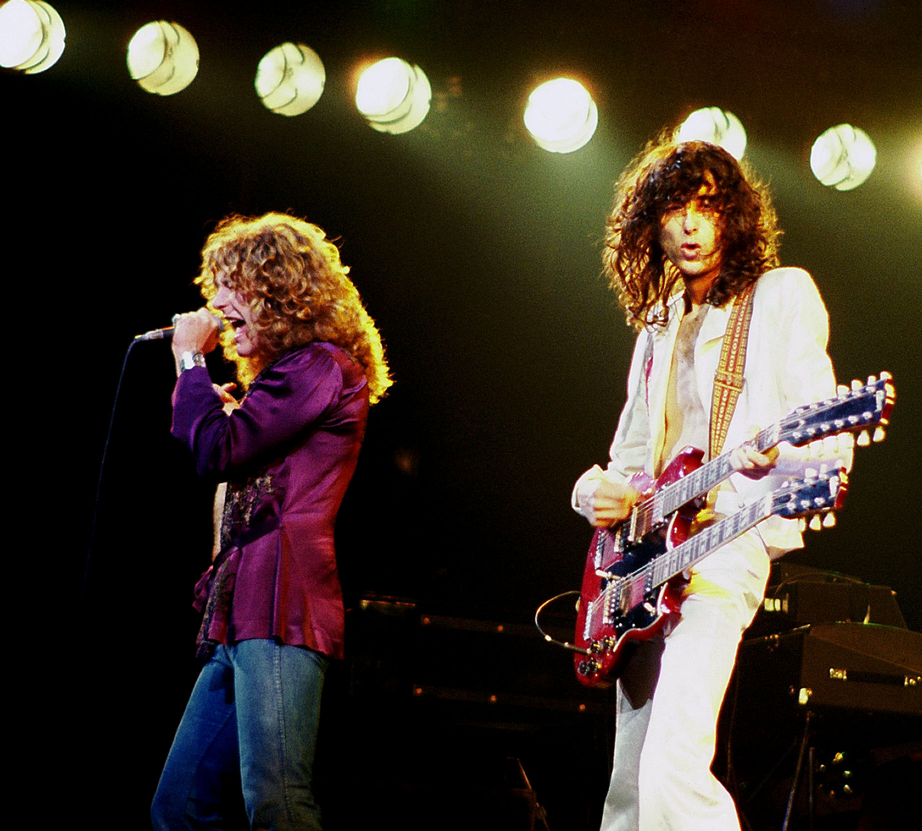 Robert Plant in a purple shirt singing into a microphone beside Jimmy Page playing a red double-neck guitar under bright stage lights, 1977.