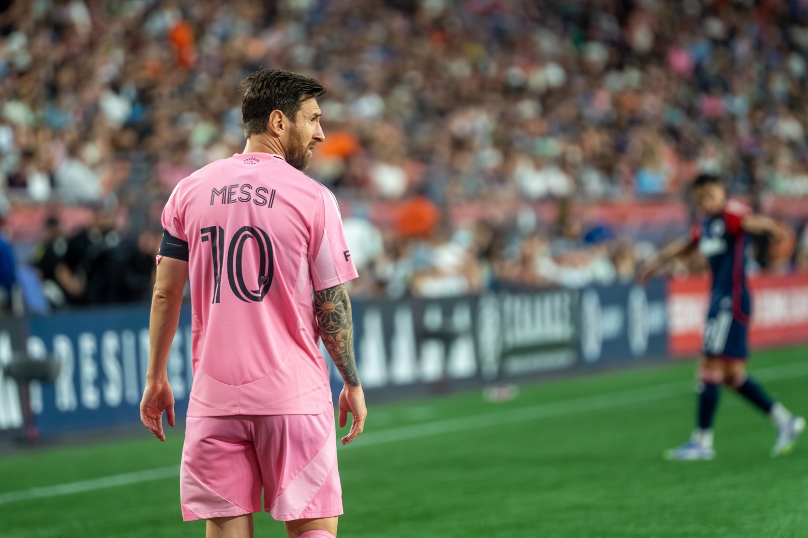 Lionel Messi in a pink Inter Miami No. 10 jersey and black shorts on the field during a match, with fans in the stands behind him.
