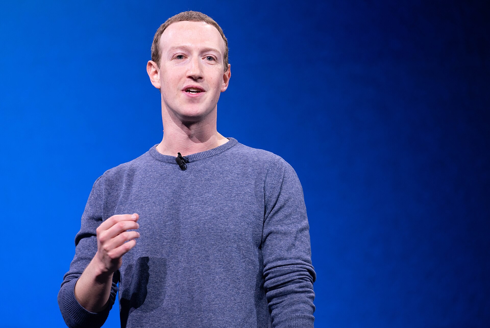 Mark Zuckerberg in a blue crewneck sweater speaking on stage against a bright blue background