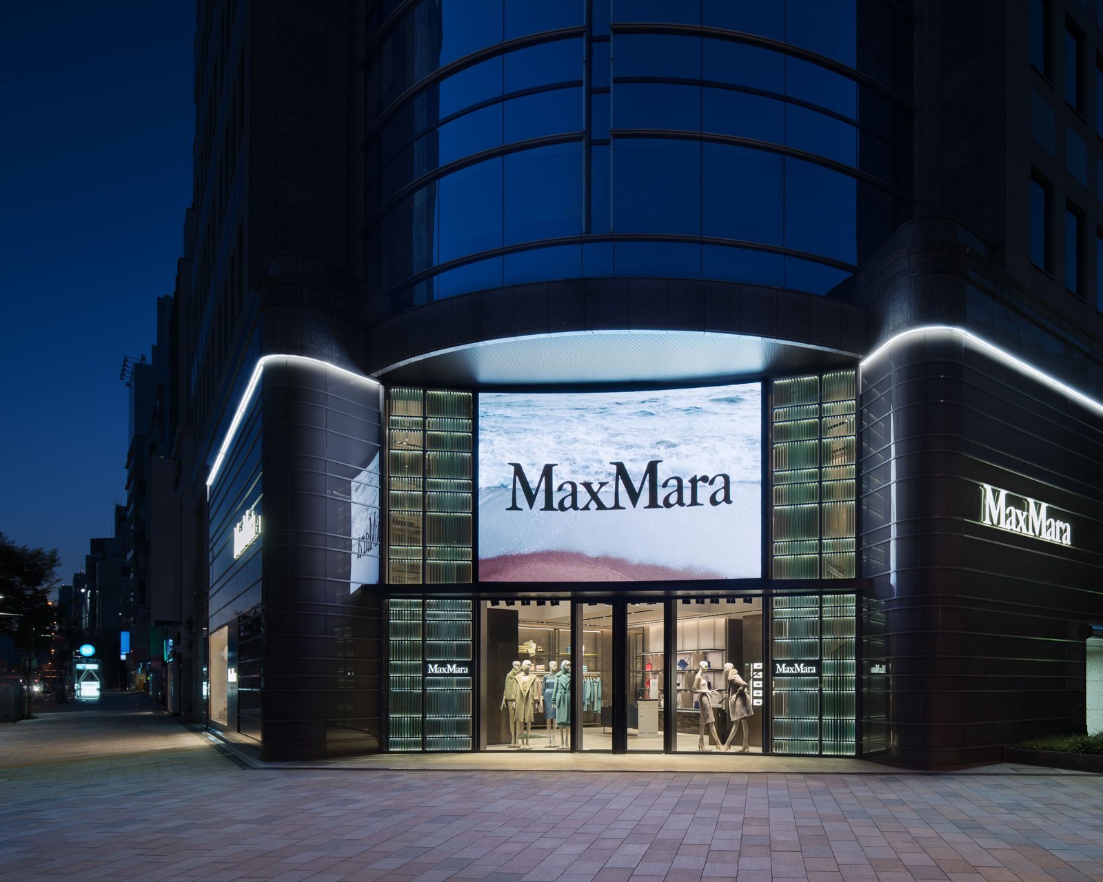 The illuminated glass exterior of a Max Mara boutique in Tokyo's Minami-Aoyama district at dusk, with mannequins dressed in coats in the windows.