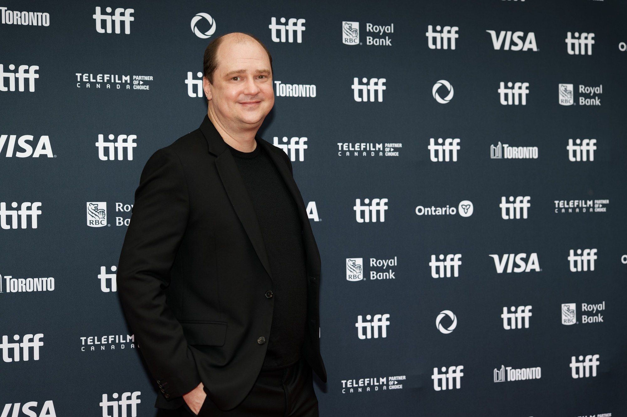 Director Mike Flanagan, wearing a black suit and black shirt, smiling on a red carpet in front of a TIFF sponsor backdrop.